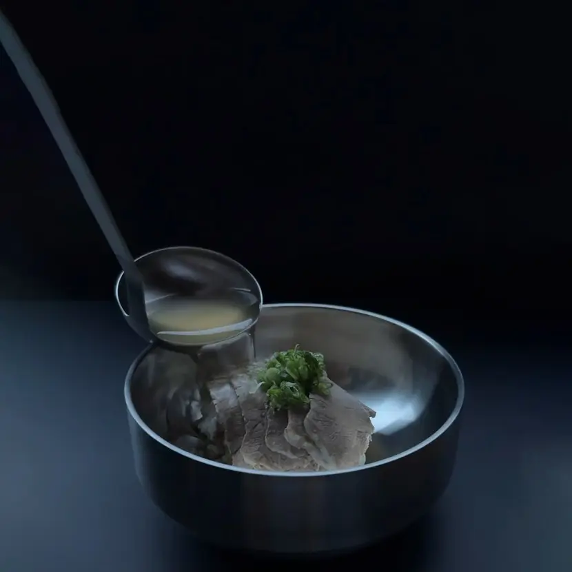 Ladle pouring broth into a metal bowl with sliced meat and chopped green onions on top.
