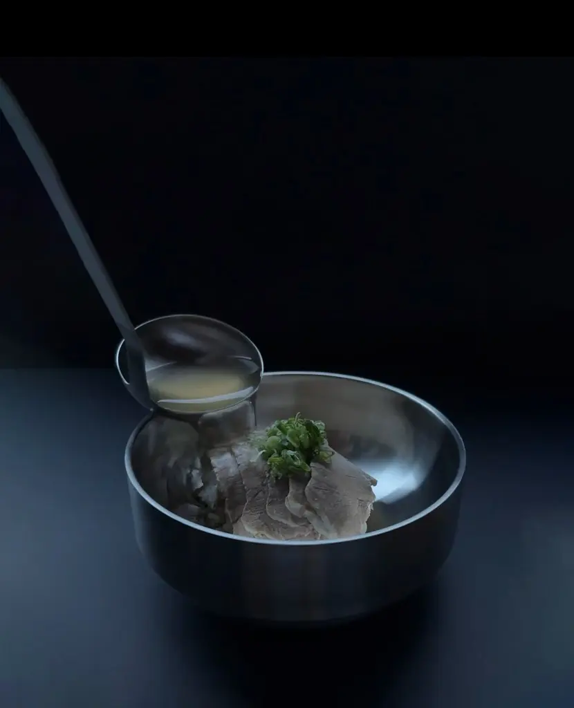Ladle pouring broth into a metal bowl with sliced meat and chopped green onions on top.