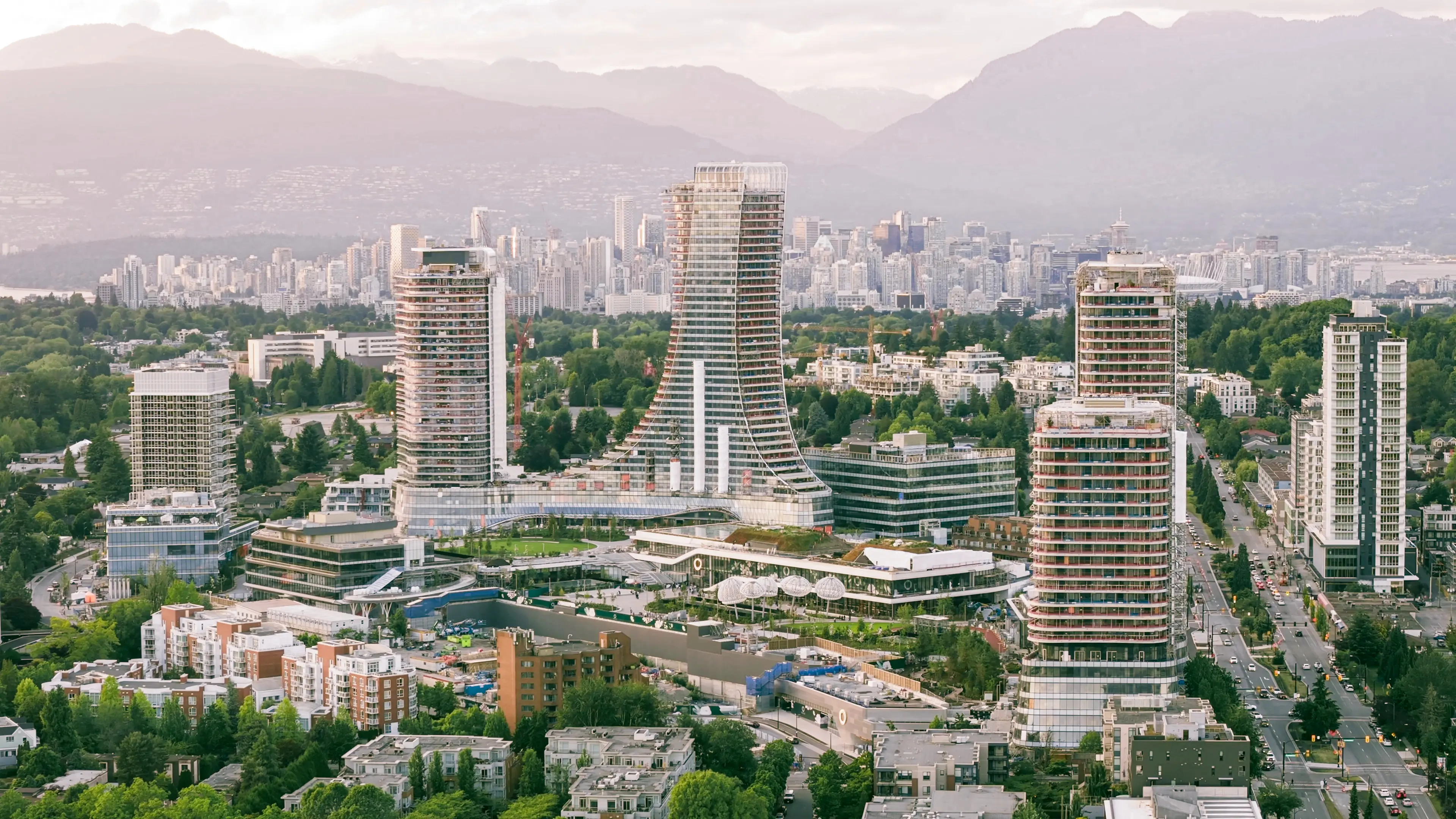 A cityscape with modern high-rise buildings surrounded by greenery and mountains in the background under a cloudy sky.