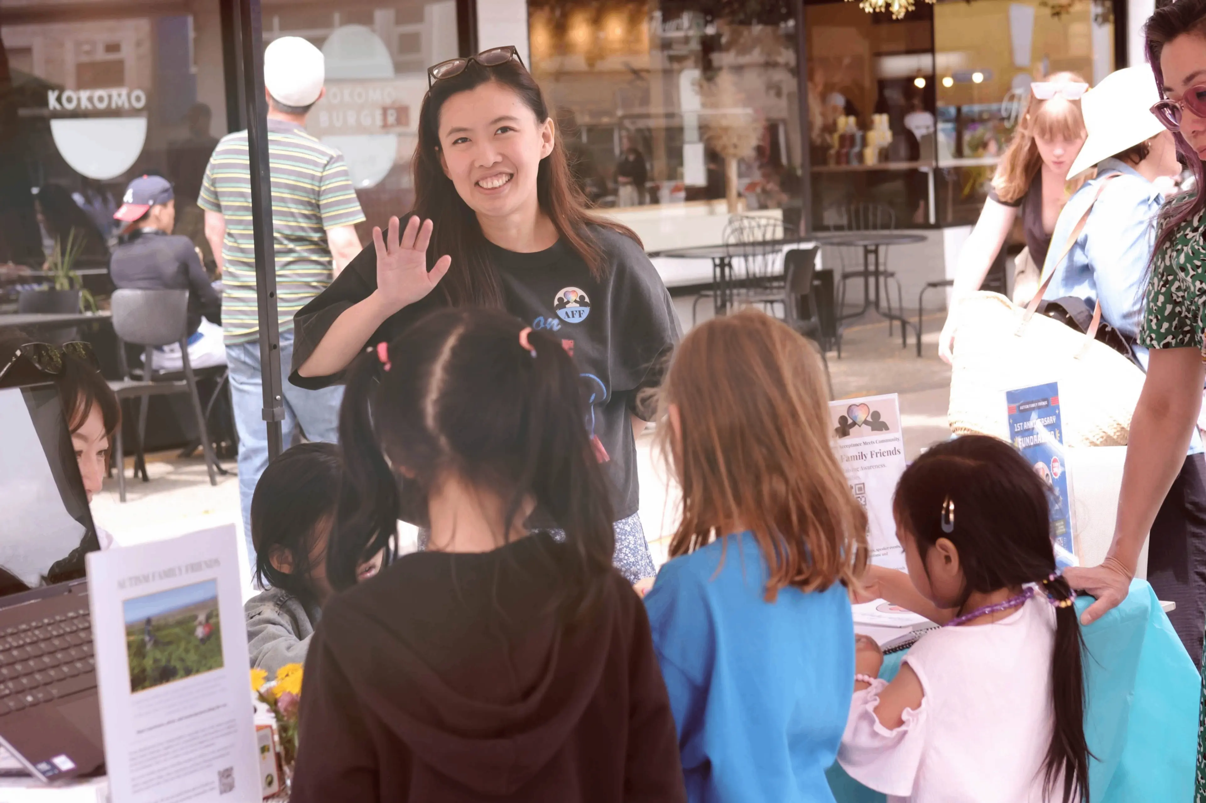 A person smiling and waving behind a table with children facing them, outdoors near a cafe named Kokomo.