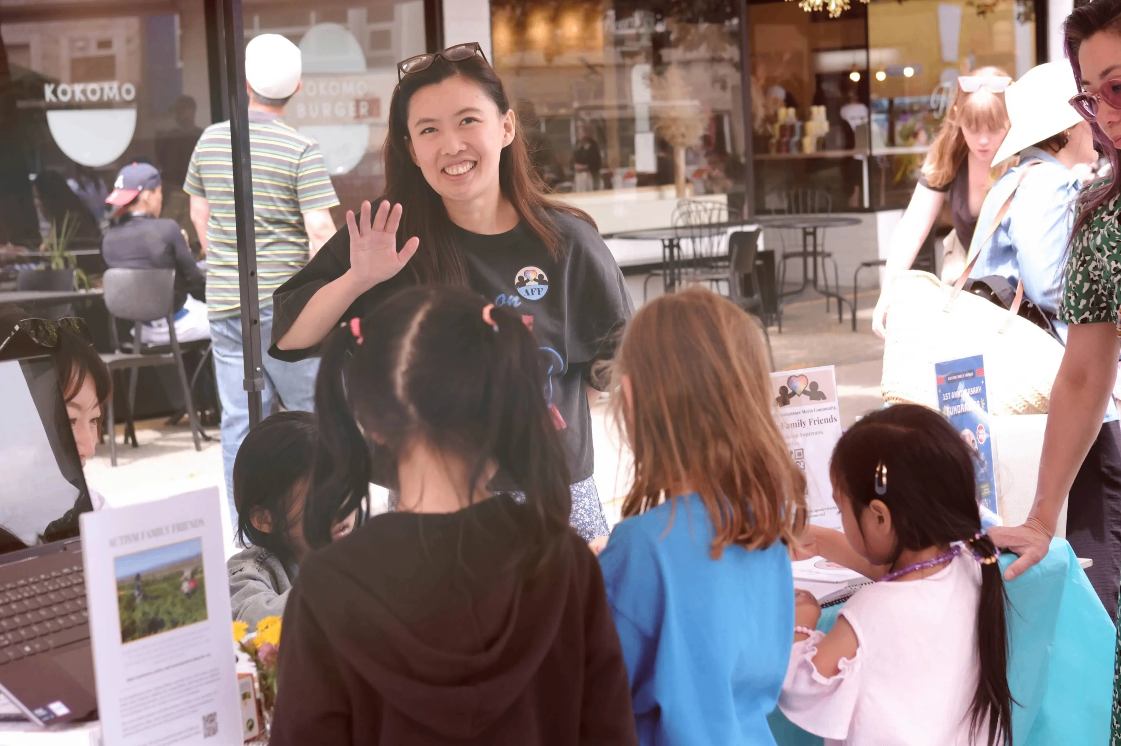 A person smiling and waving behind a table with children facing them, outdoors near a cafe named Kokomo.