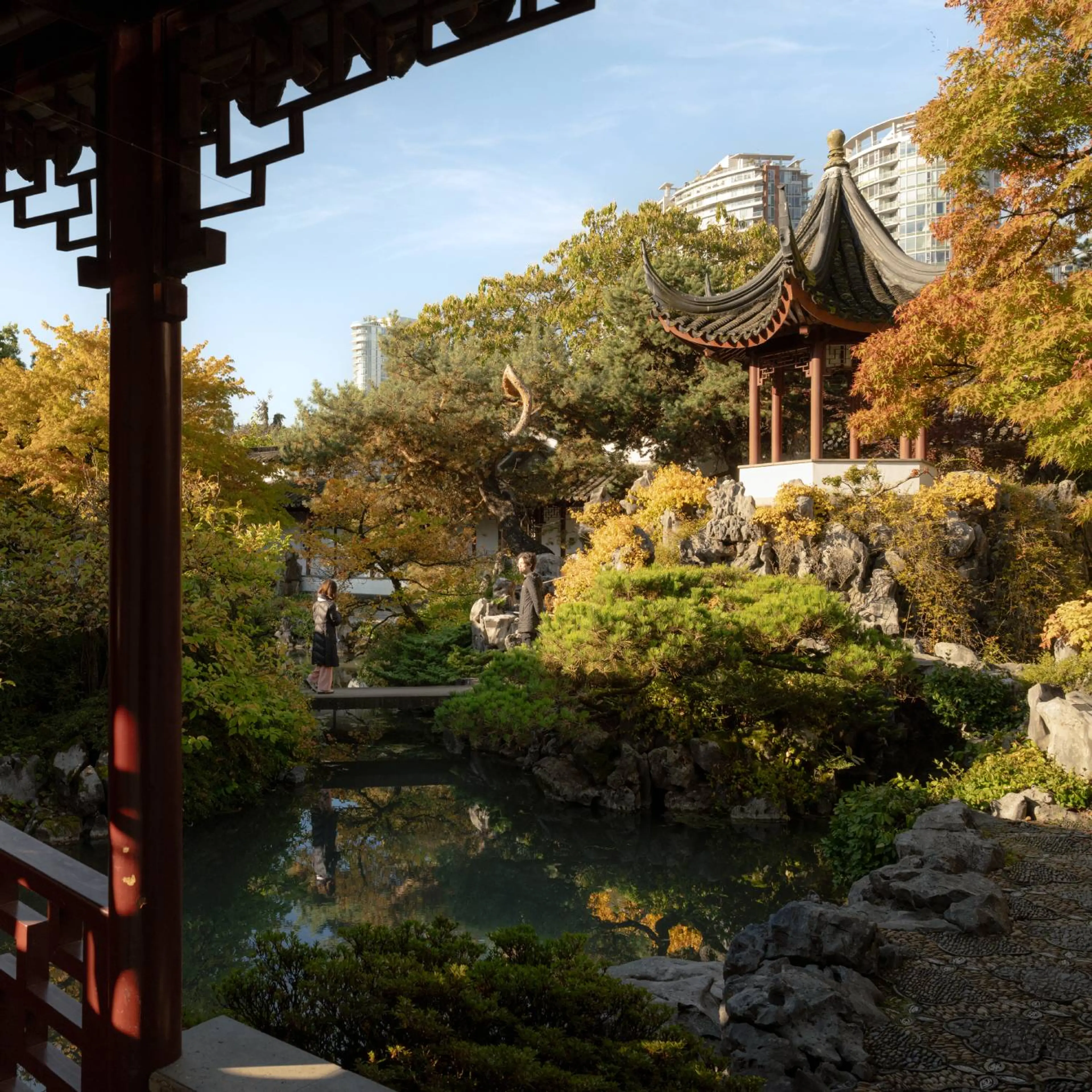 Two people standing on a small bridge over a pond in a garden with autumn foliage and a traditional Asian pavilion.