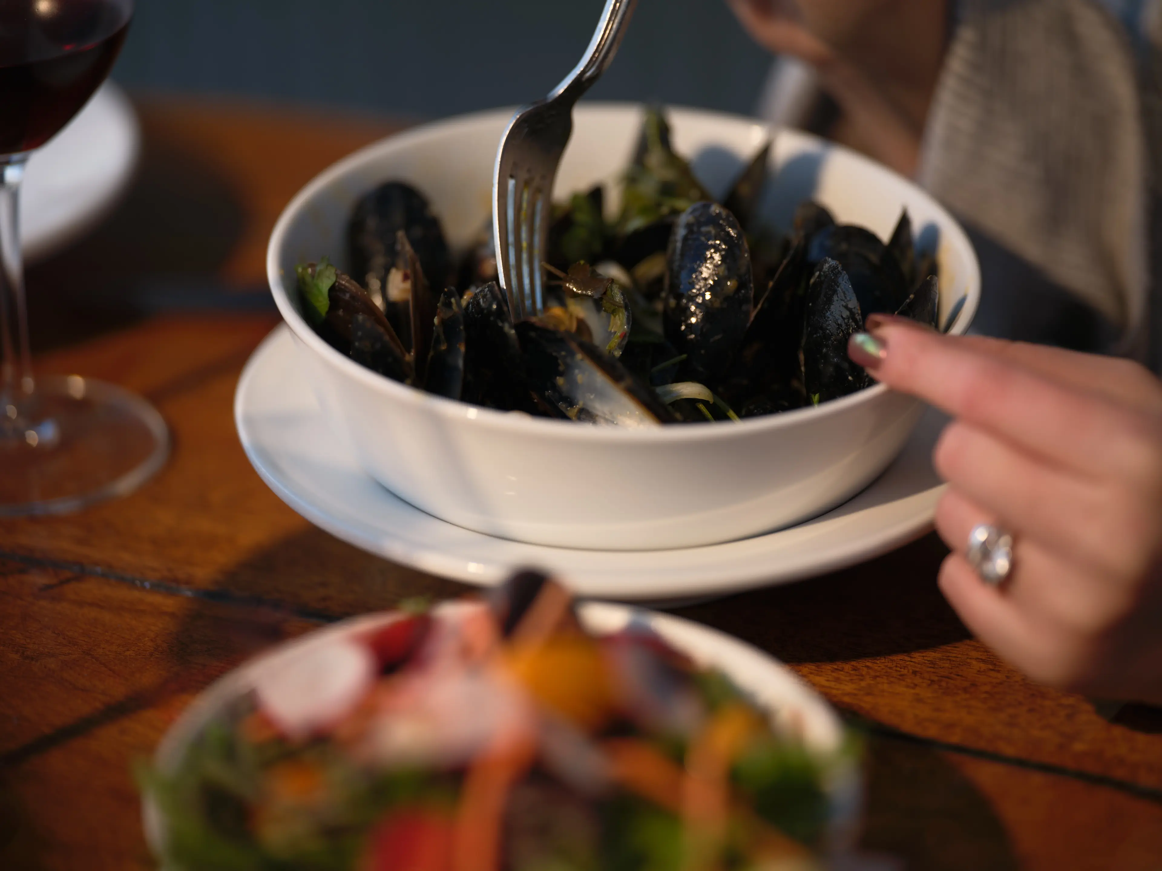 Person using a fork to eat mussels from a white bowl on a wooden table with a glass of red wine and a salad bowl nearby.