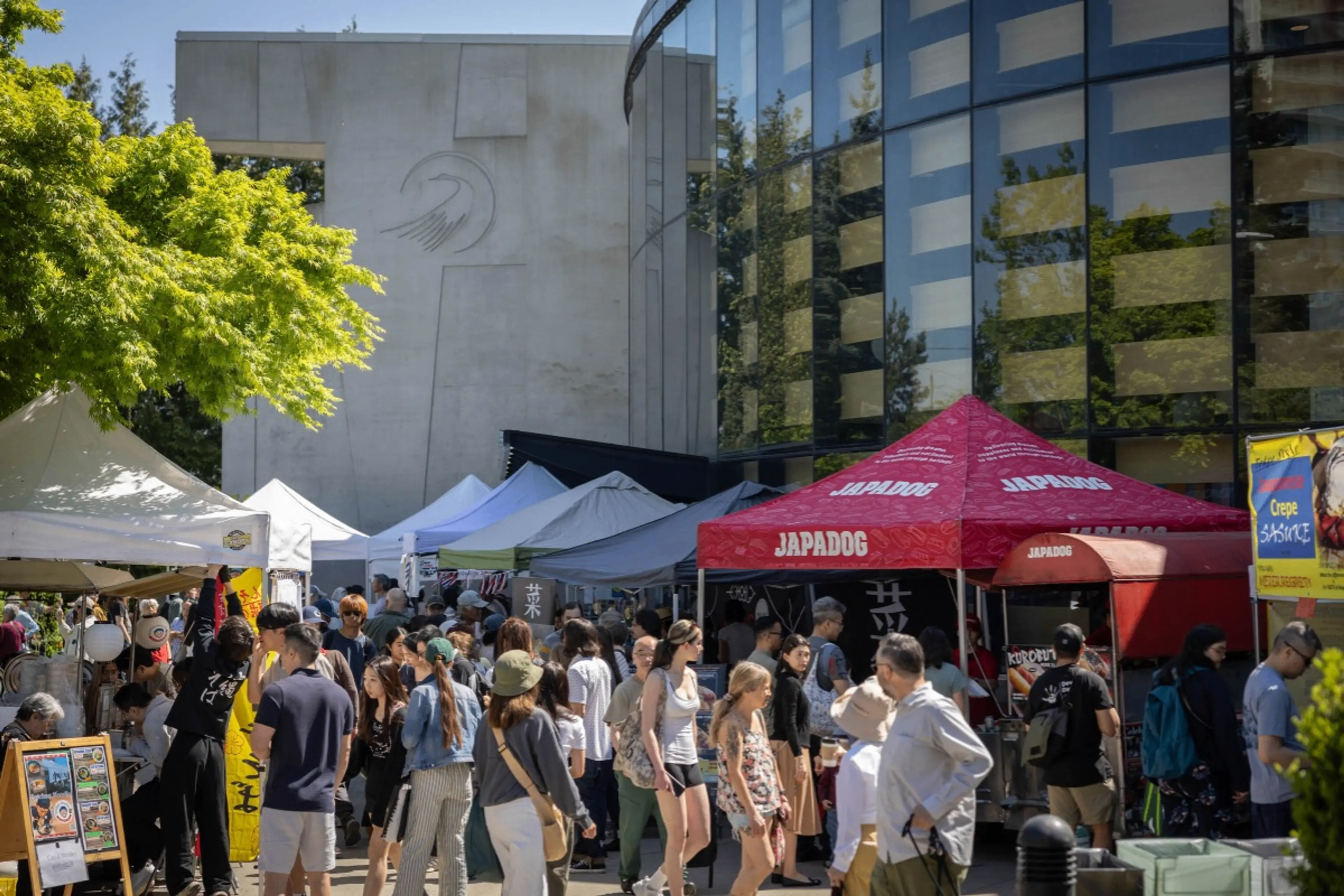 Crowd of people walking and standing near outdoor market tents, including a red Japadog tent, on a sunny day.