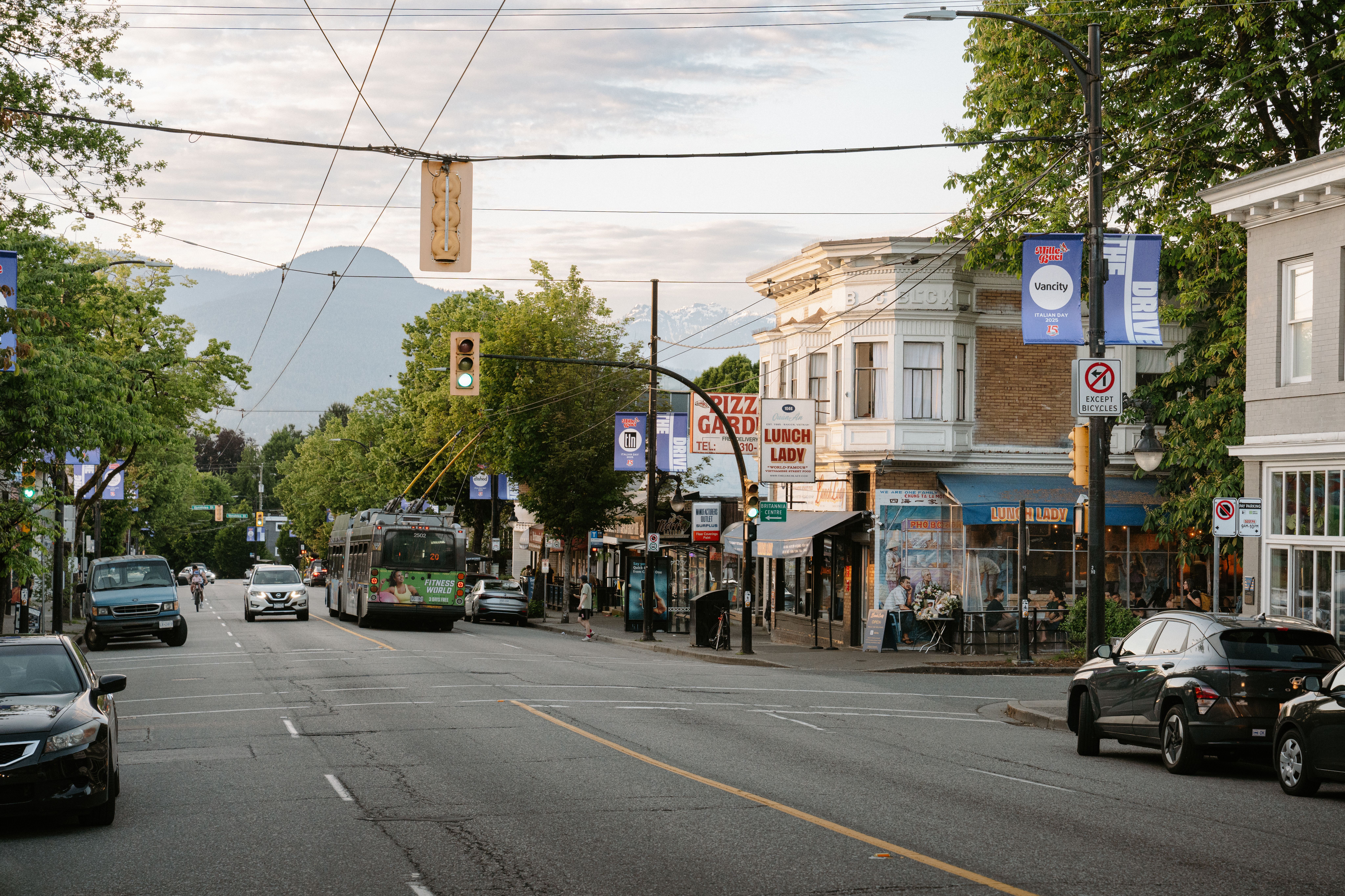Street view with cars, a bus, cyclists, and a building with signs for Pizza Garden and Lunch Lady under a cloudy sky.