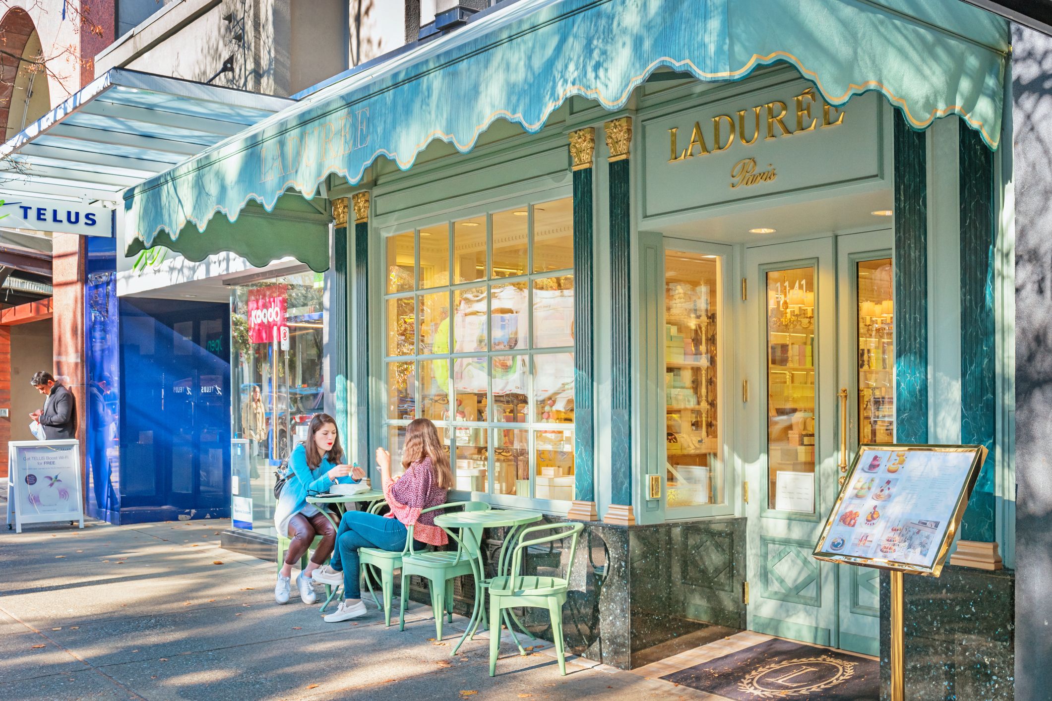 Two people sitting at a green outdoor table with chairs outside a pastel green and gold Ladurée Paris storefront on a sunny day.