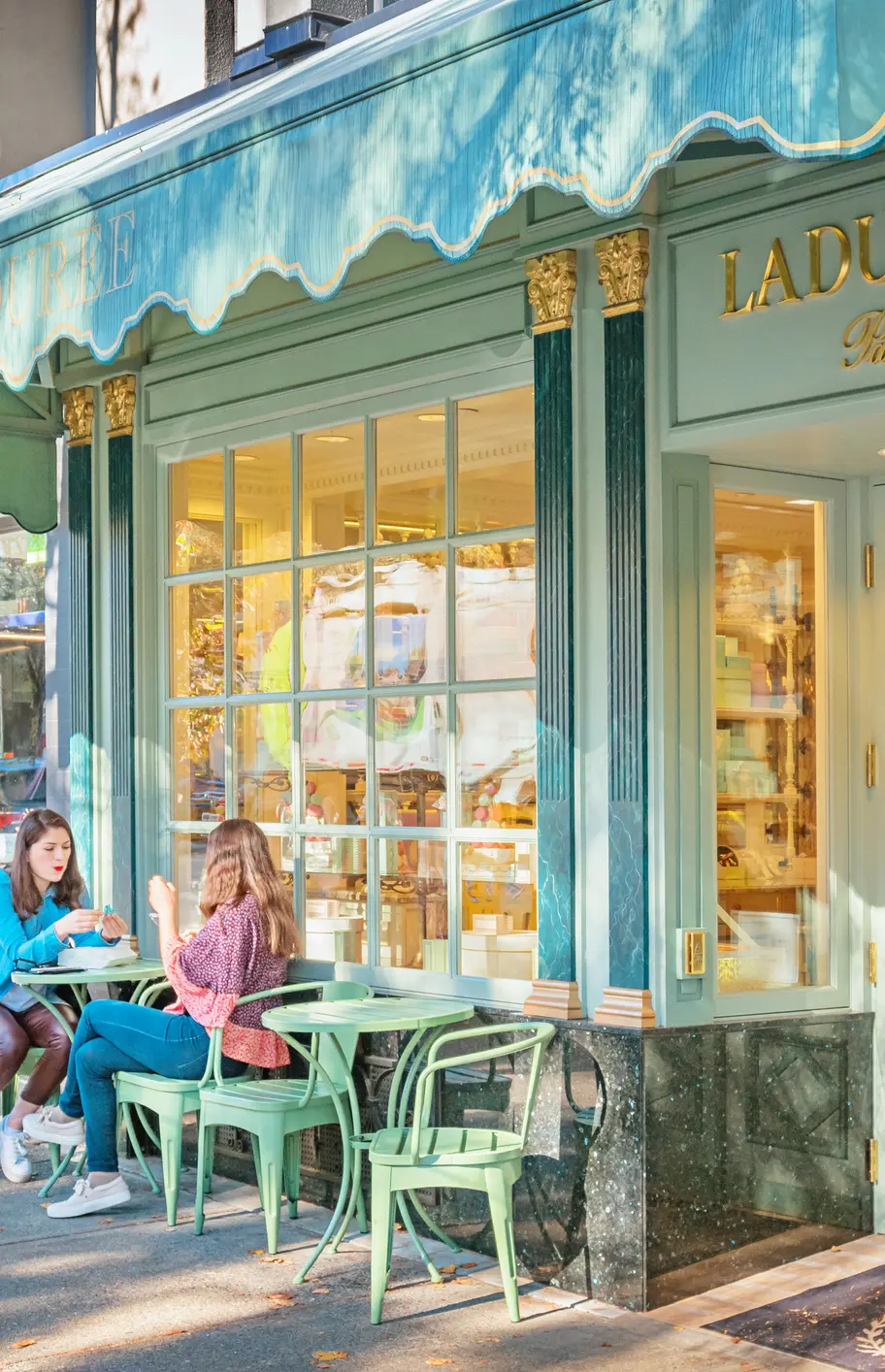 Two people sitting at a green outdoor table with chairs outside a pastel green and gold Ladurée Paris storefront on a sunny day.