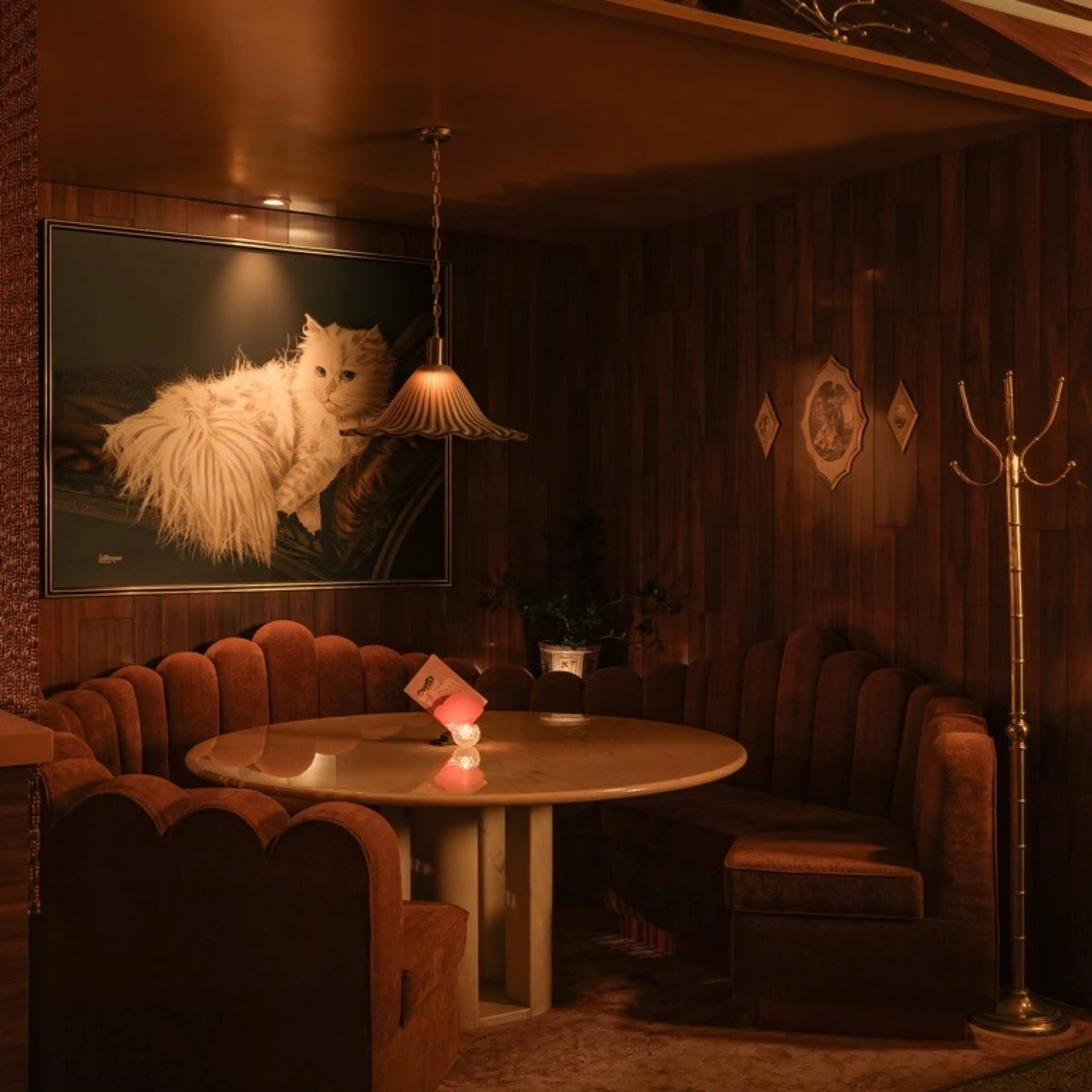 Cozy corner seating with brown velvet chairs around a round table under a hanging lamp, wood-paneled walls, and a large painting of a fluffy white cat.