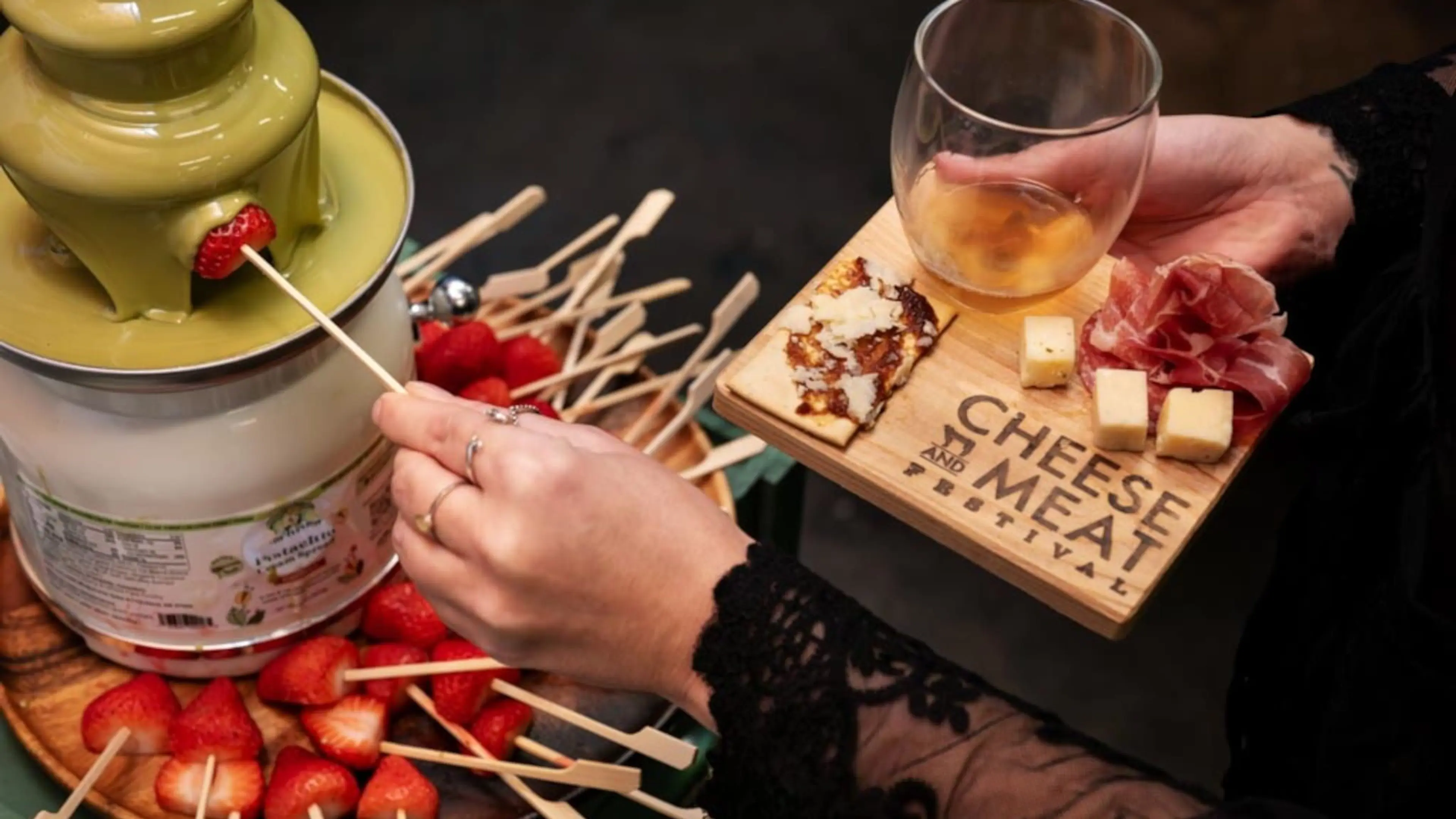 Person dipping a strawberry on a skewer into a green chocolate fountain, holding a wooden board with cheese, meat, and a glass of drink at a Cheese and Meat Festival.