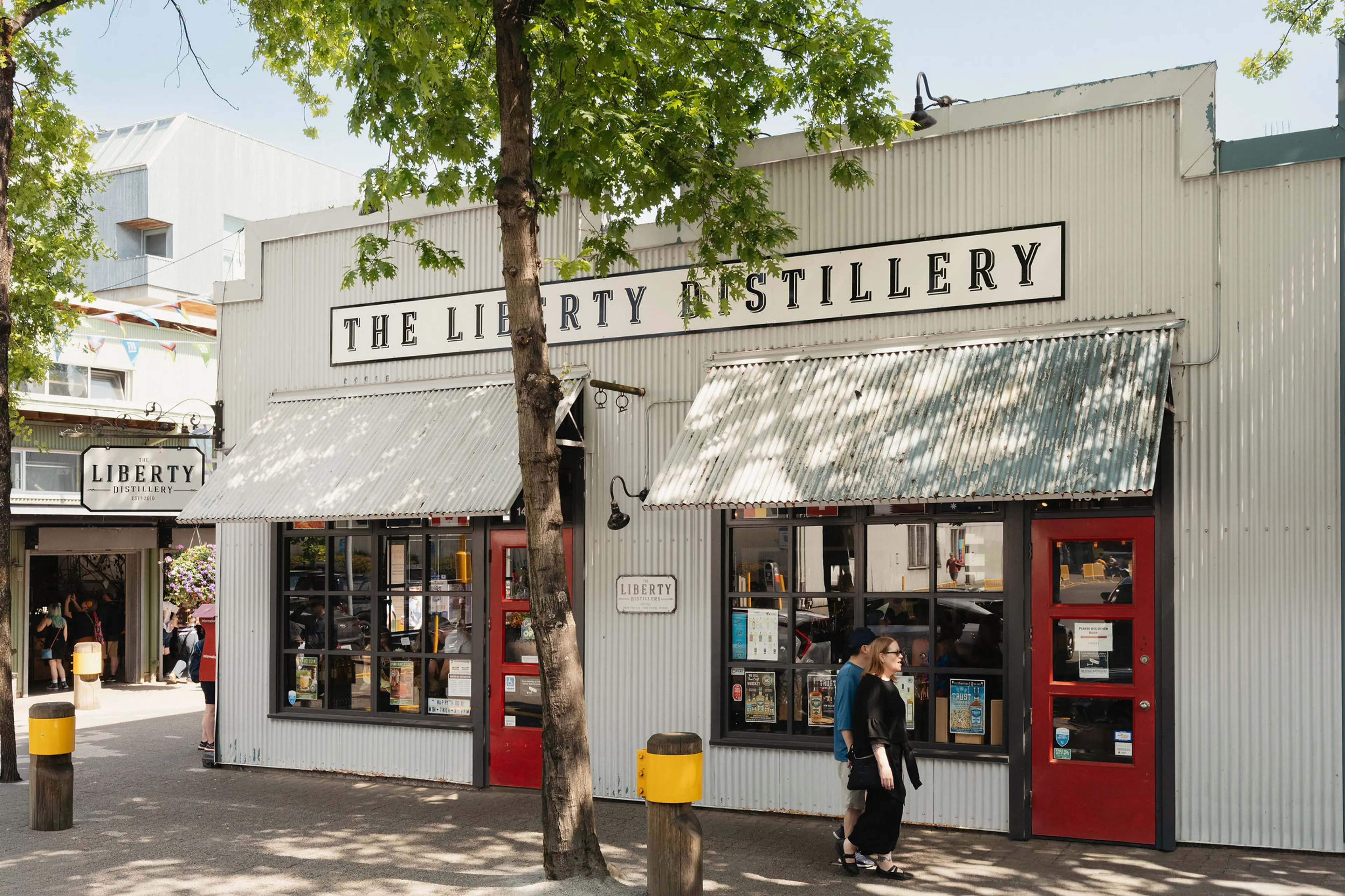 Exterior of The Liberty Distillery with two people walking past red doors and large windows under metal awnings.