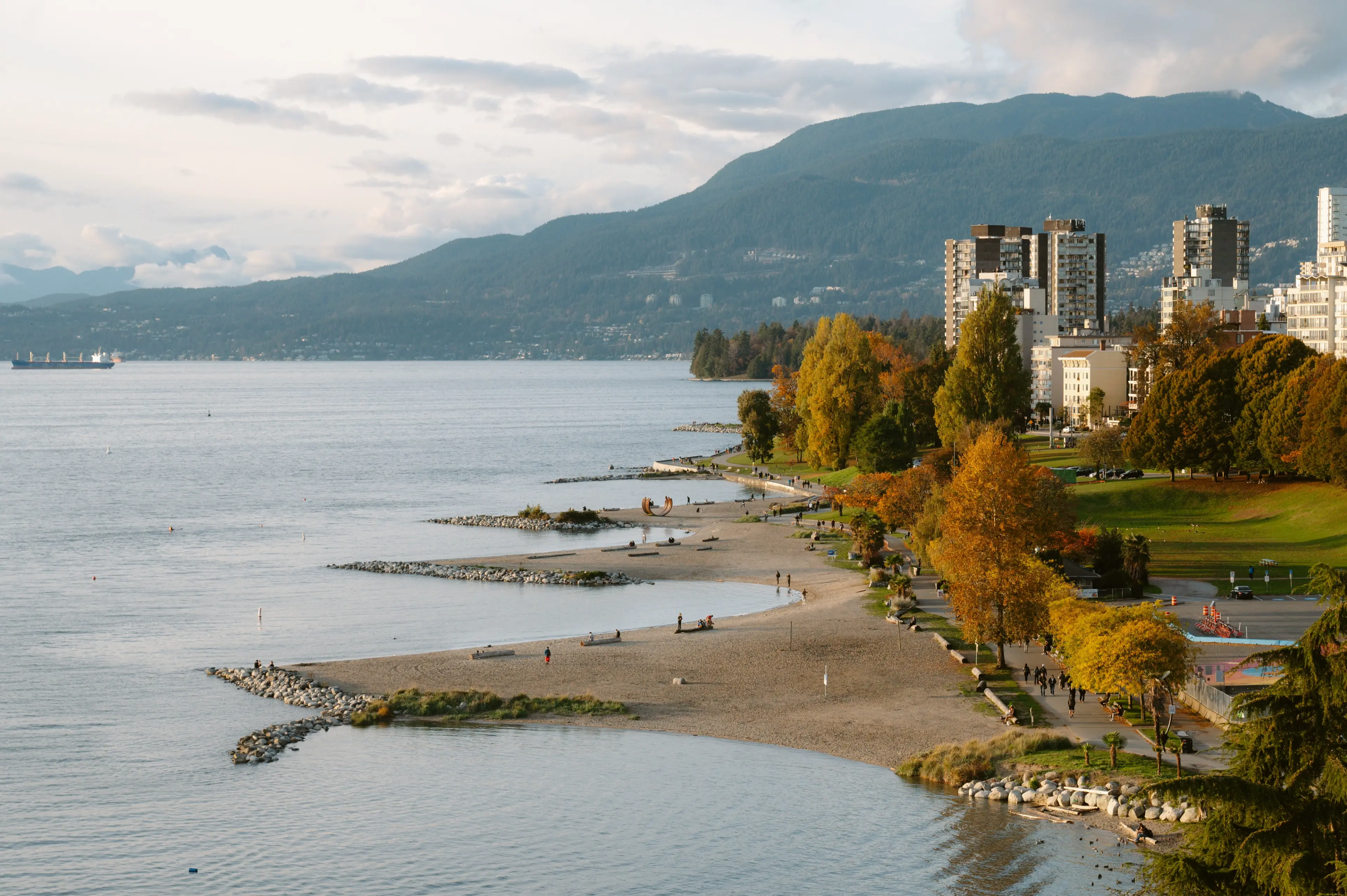 Aerial view of English Bay framed by autumn-colored trees, with the North Shore mountains in the background.