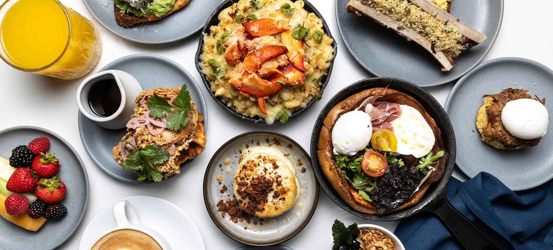 Assorted breakfast dishes including fruit, toast with greens, mac and cheese with lobster, eggs, coffee, and orange juice on a white table.