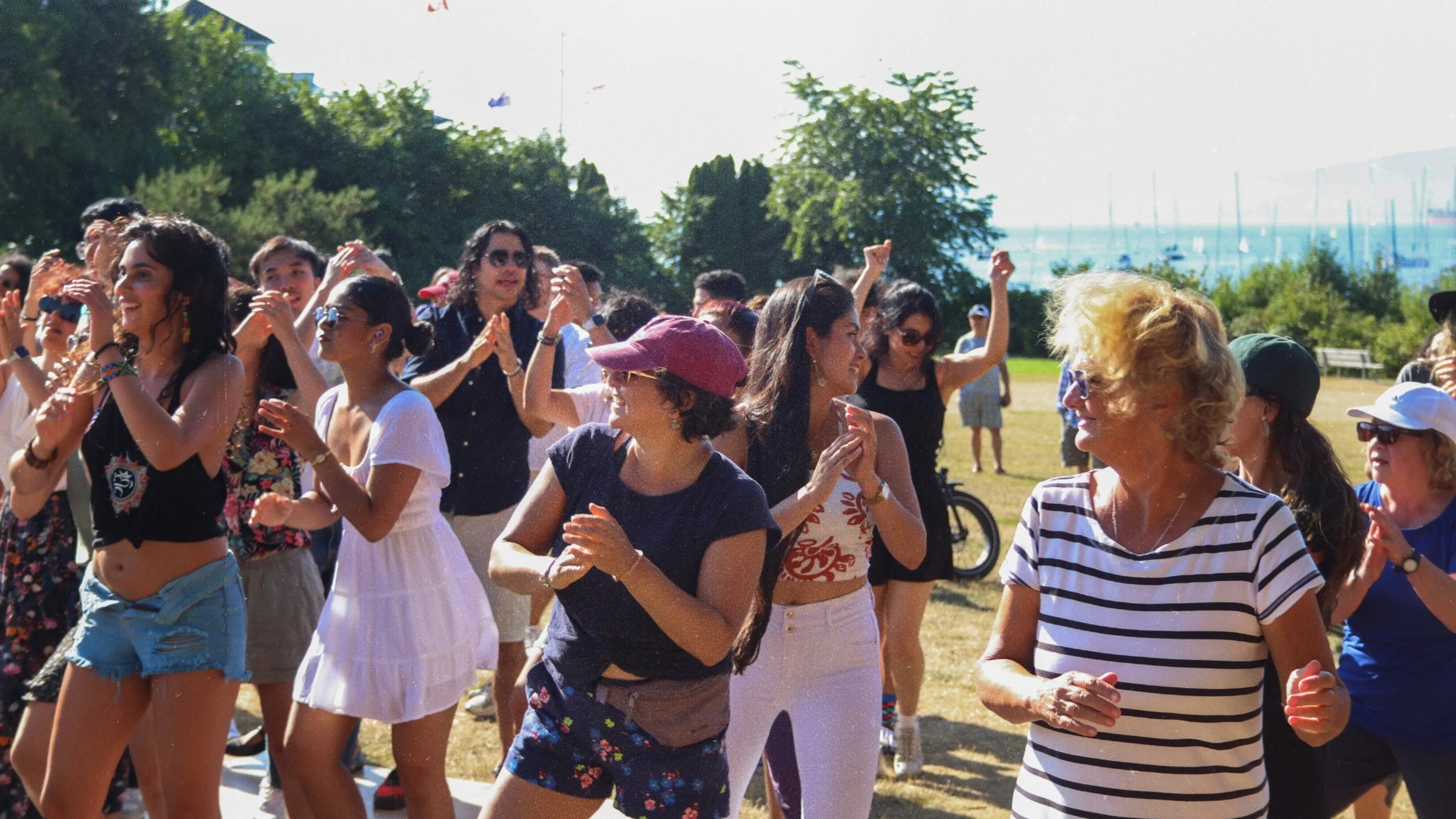 A diverse group of people dancing outdoors on a sunny day in a park with trees and water in the background.