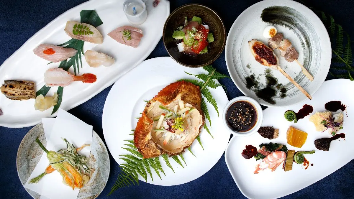 Assorted gourmet dishes including sushi, skewers, tempura, seafood, and sauces arranged on white plates over a dark blue surface.