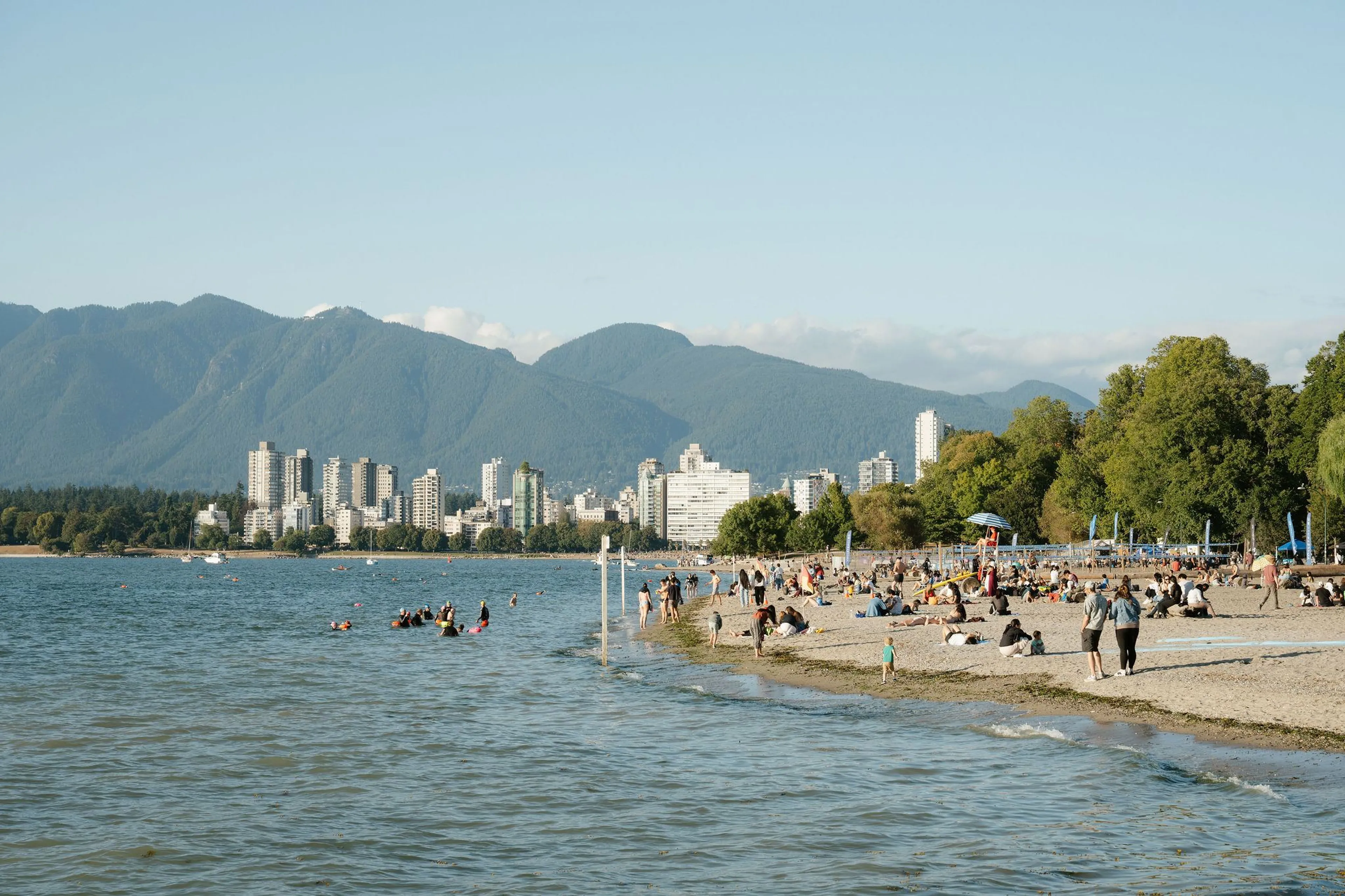Crowded beach with people swimming and relaxing, city skyline and forested mountains in the background under clear sky.