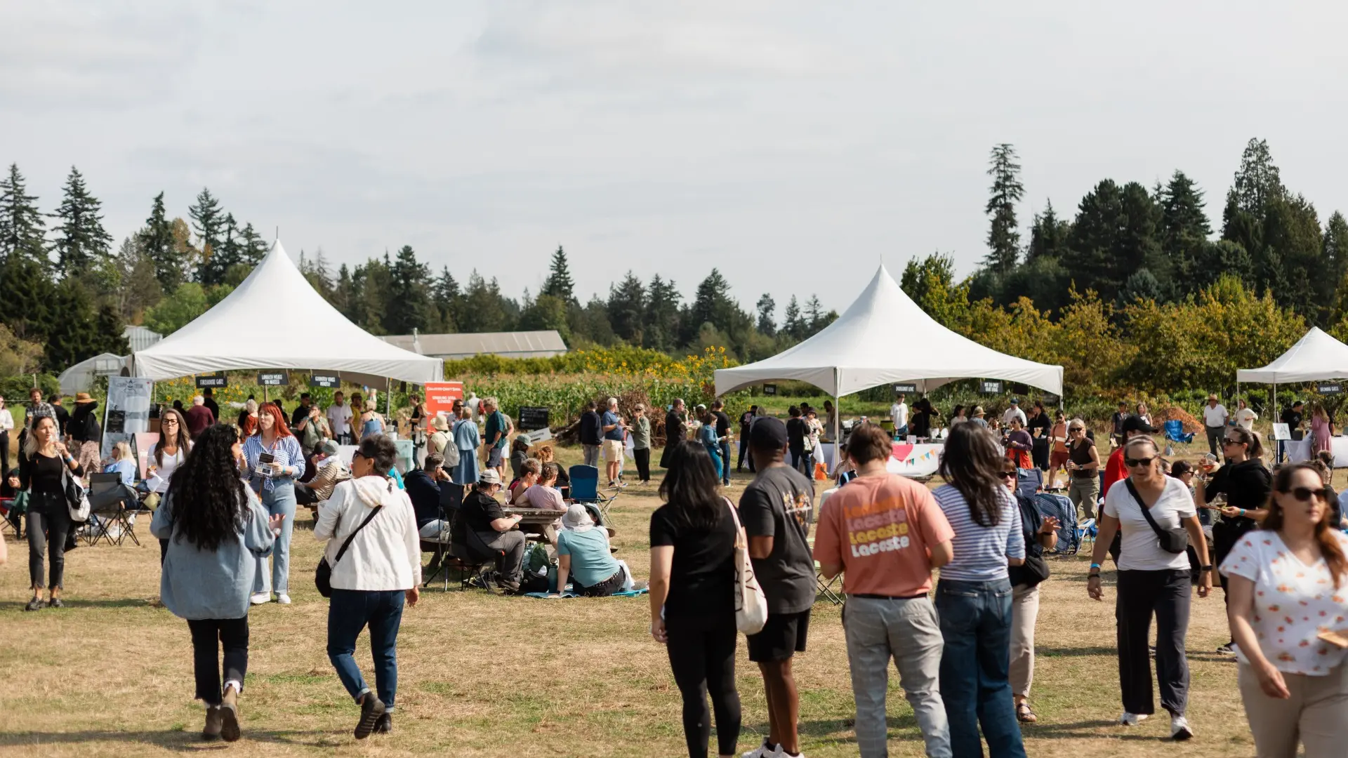 Crowd of people walking and sitting on grass near white event tents in an outdoor park with trees in the background.