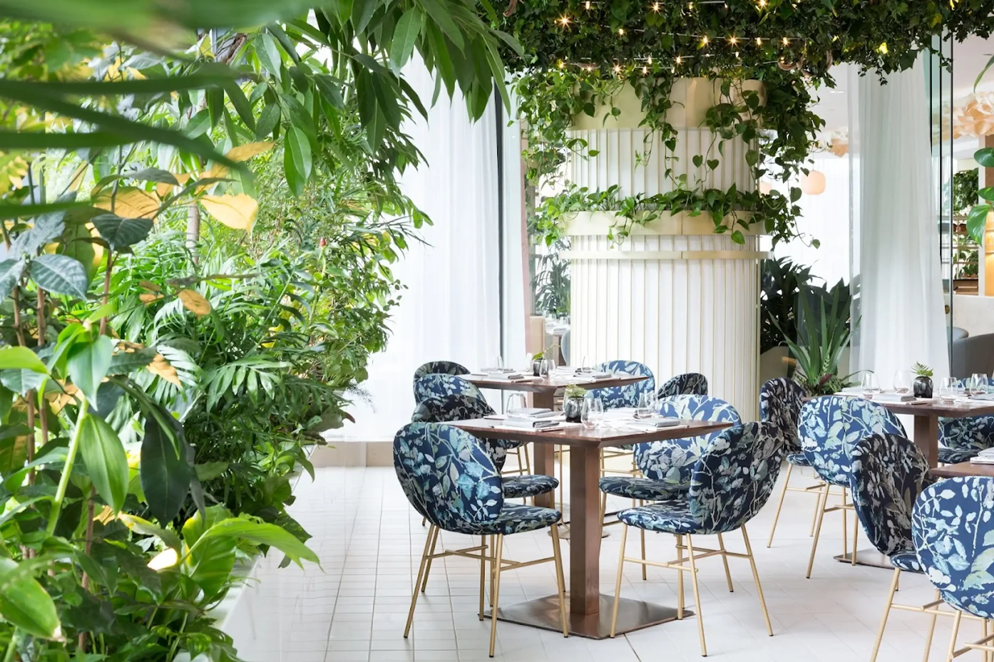 Indoor dining area with tables and floral chairs surrounded by lush plants and curtains at the Botanist restaurant in Vancouver.