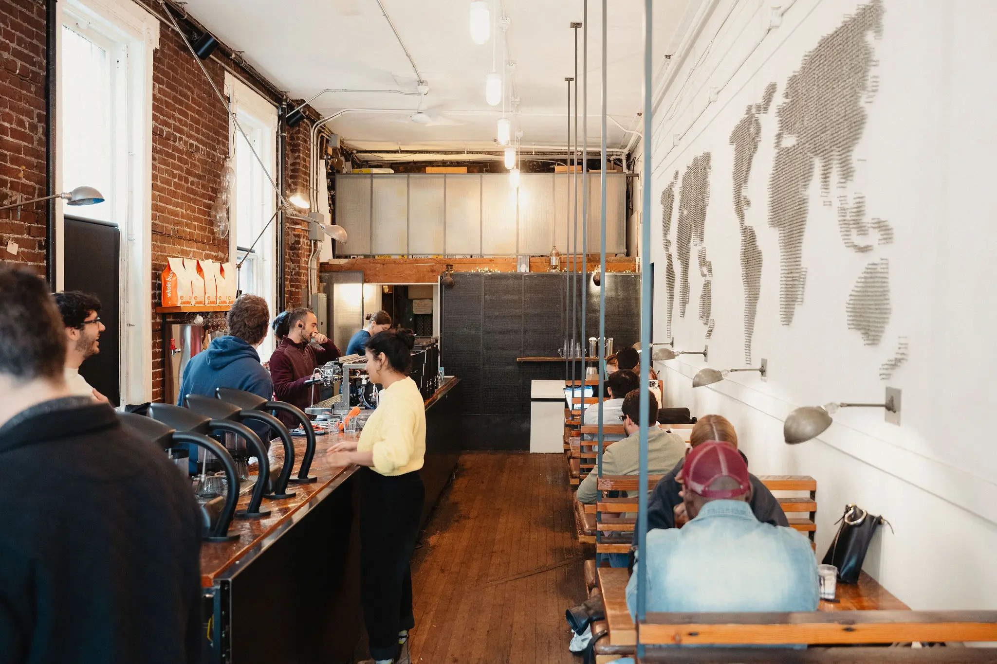 People sitting at wooden tables and a counter in a coffee shop with a world map art on the white wall.