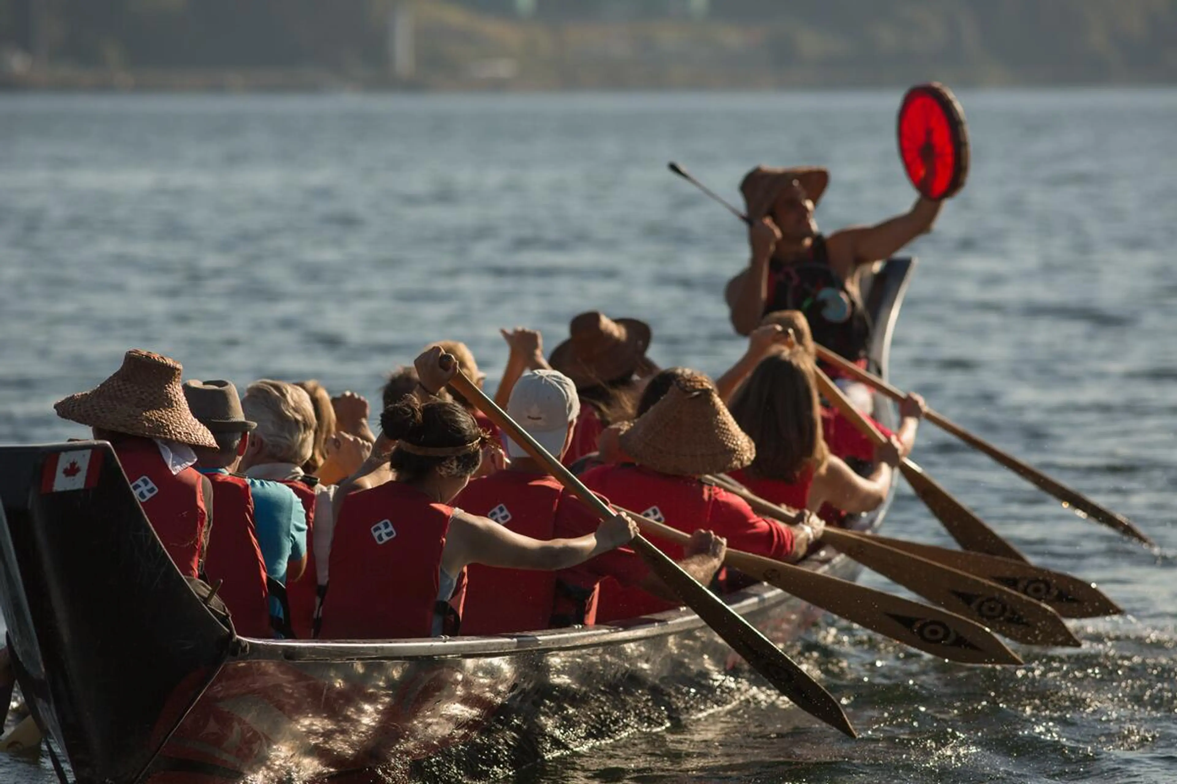 A group of people paddling a canoe on a body of water, with one person at the front holding a red drum.