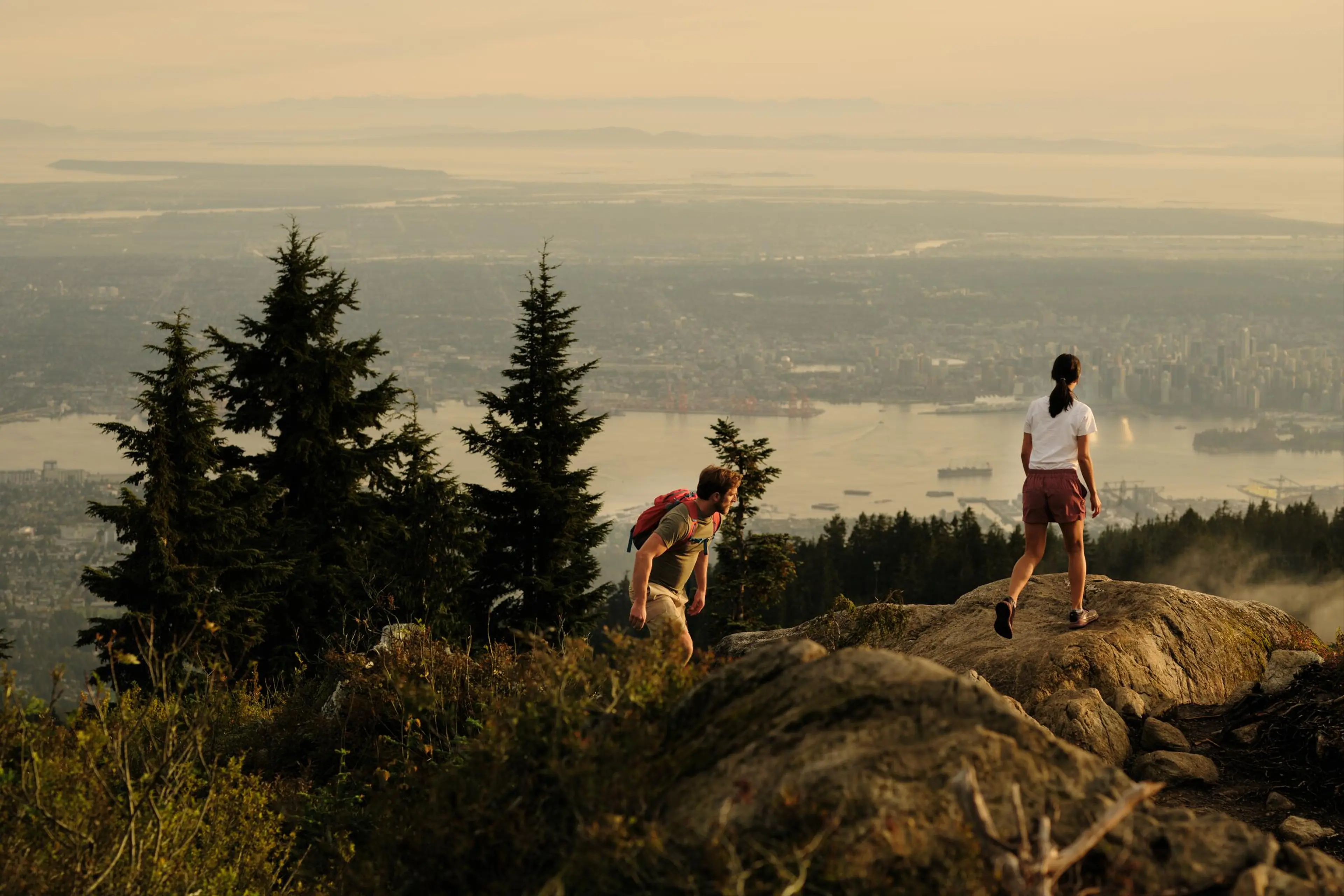 Two hikers on a rocky mountain trail overlooking a city and water at sunset.