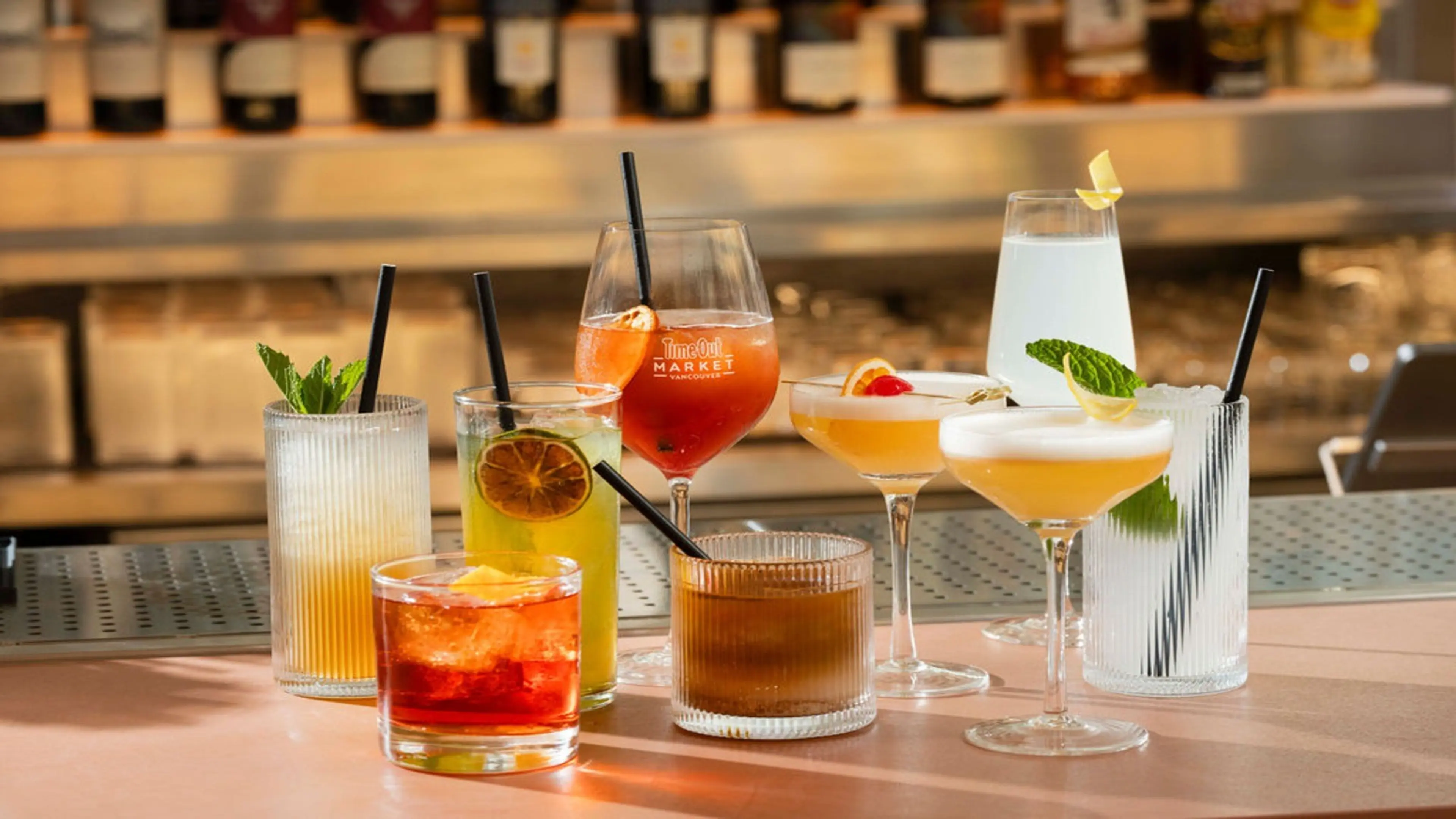 Eight colorful cocktails in various glasses with garnishes and straws on a bar counter with bottles in the background.