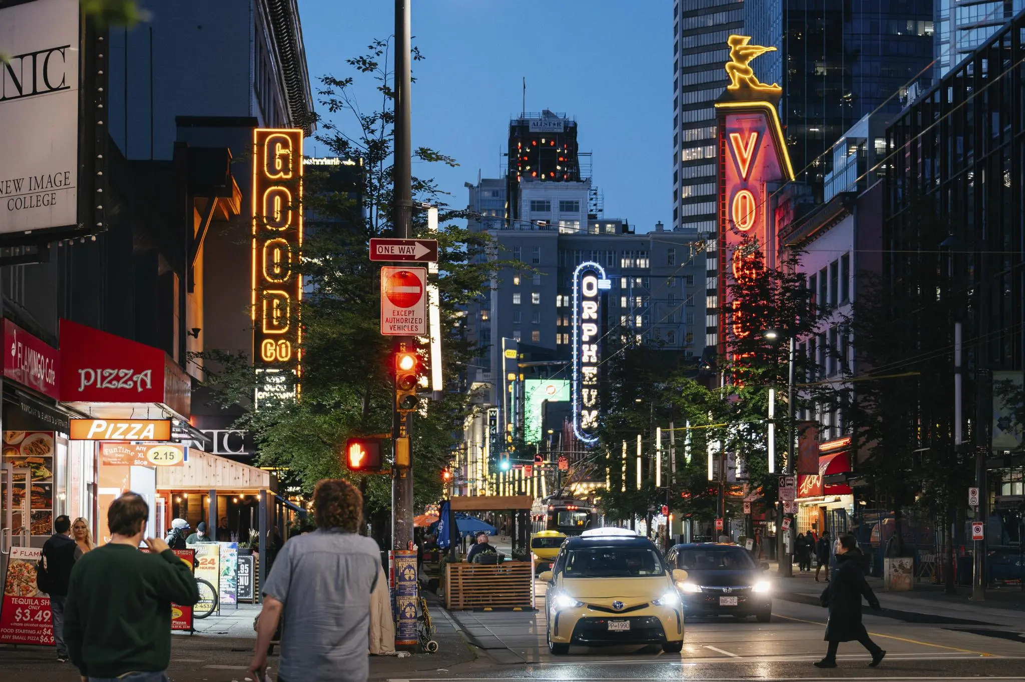 City street at dusk with illuminated signs reading GOOD, ORPHEUM, and VO, cars, and pedestrians crossing.