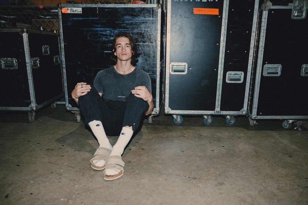Person sitting on the floor with knees up, wearing a dark shirt, black pants, white socks, and beige sandals, against black equipment cases.