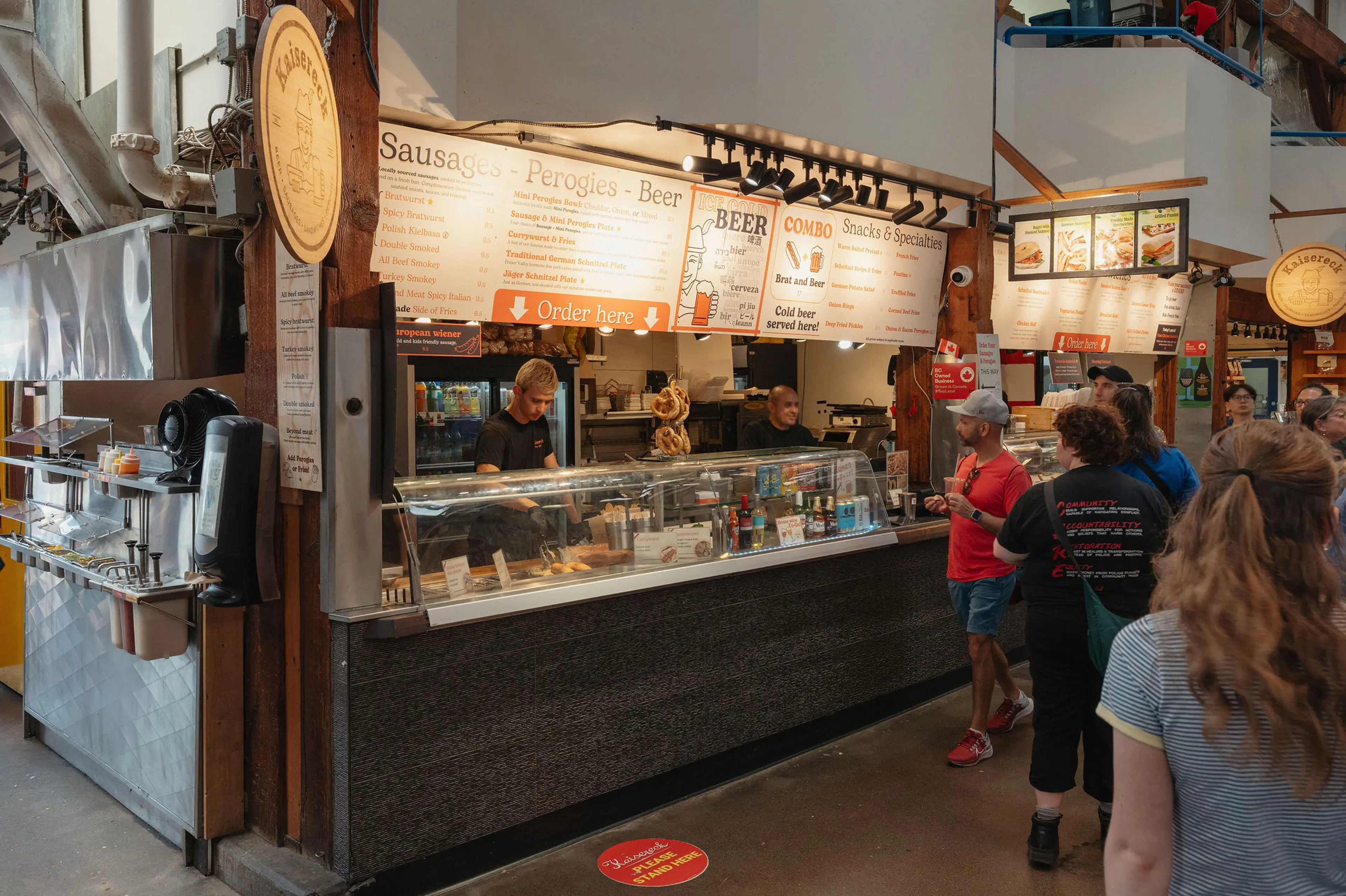 People standing in line at a food counter with menus displaying sausages, perogies, beer, and snacks in a market setting.