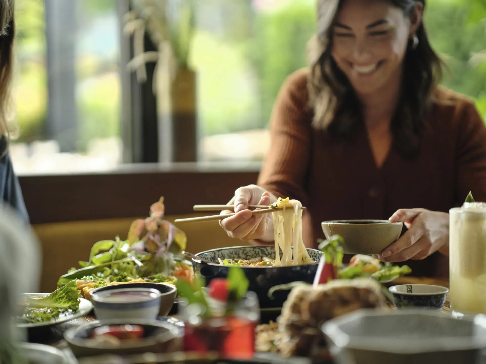 Smiling woman using chopsticks to lift noodles from a bowl at a table with various dishes and drinks.