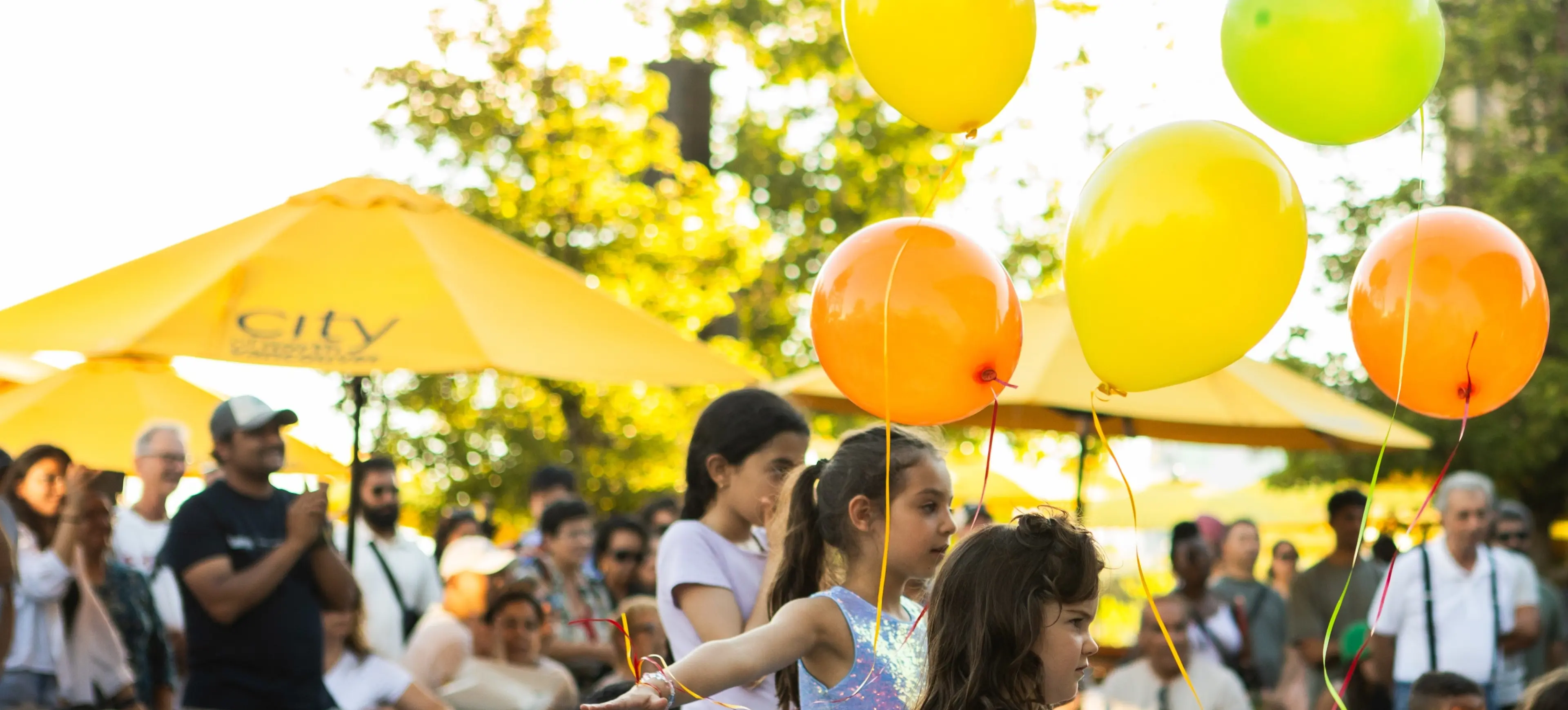 Children holding yellow, orange, and green balloons at an outdoor event with a crowd and yellow umbrellas in the background.