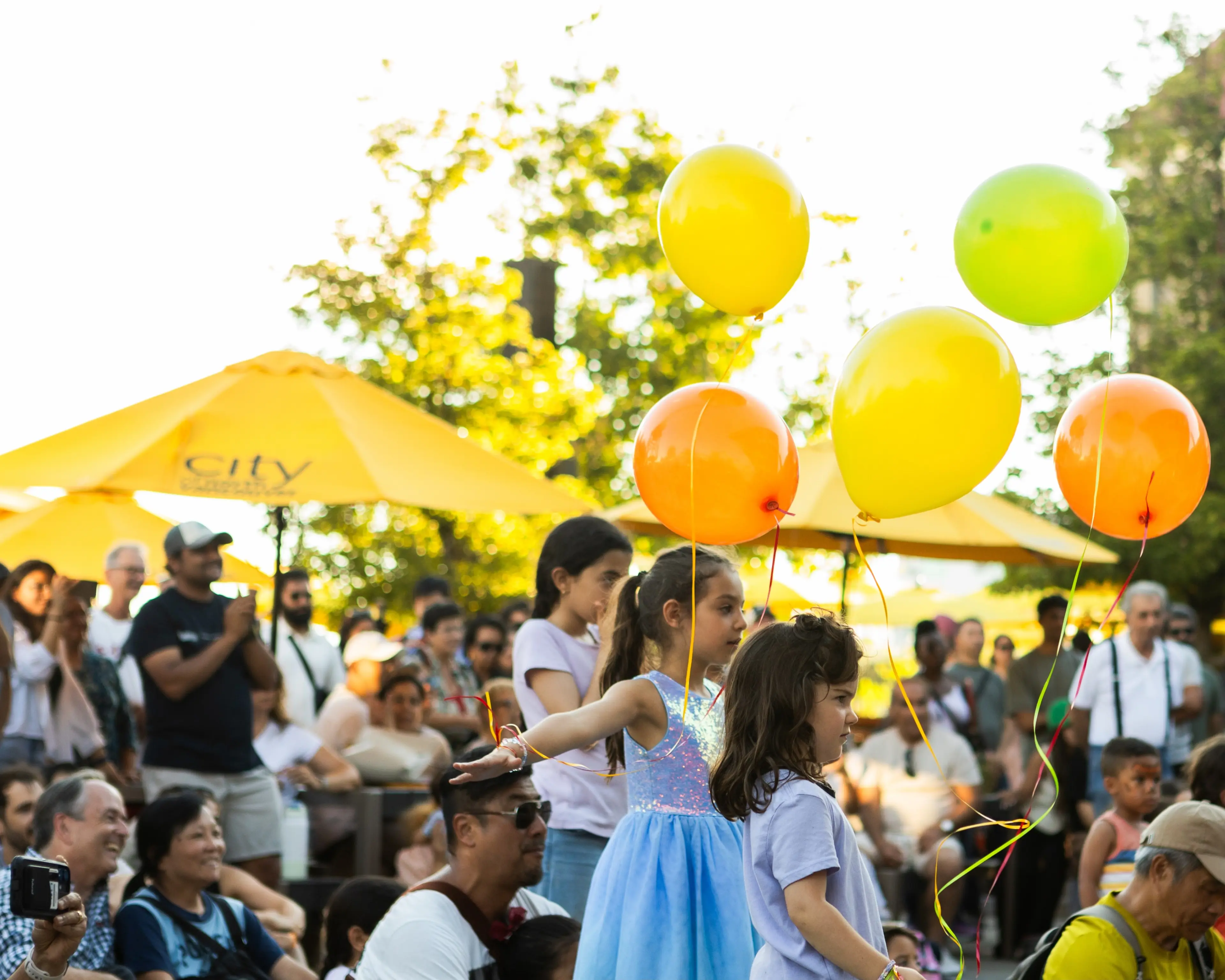 Children holding yellow, orange, and green balloons at an outdoor event with a crowd and yellow umbrellas in the background.