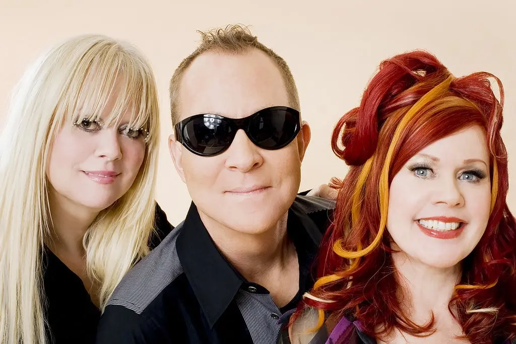 Three people posing closely, one with long blonde hair, one wearing black sunglasses, and one with red and orange styled hair smiling.
