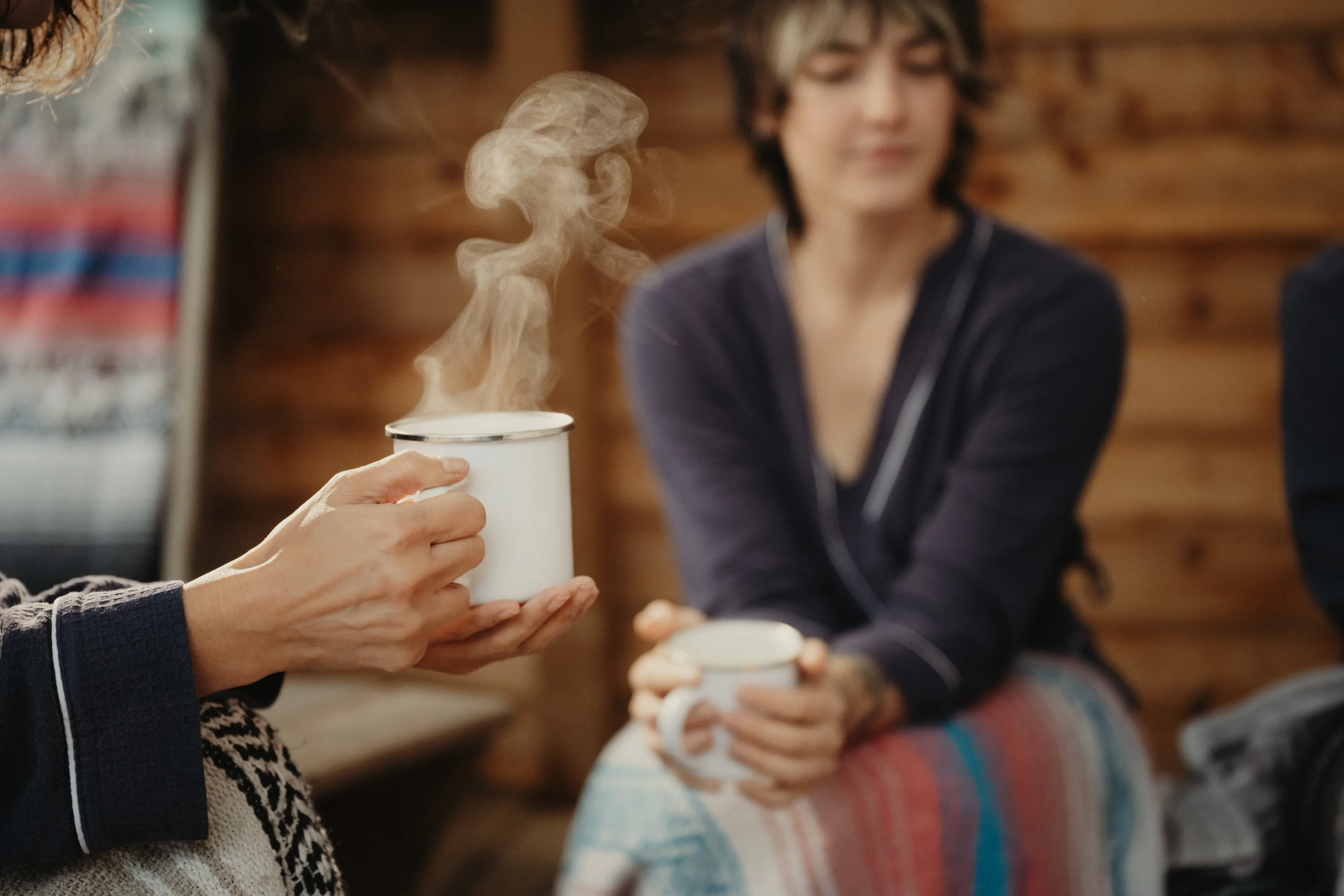 Two people sitting indoors holding steaming white mugs, one person is blurred in the background.