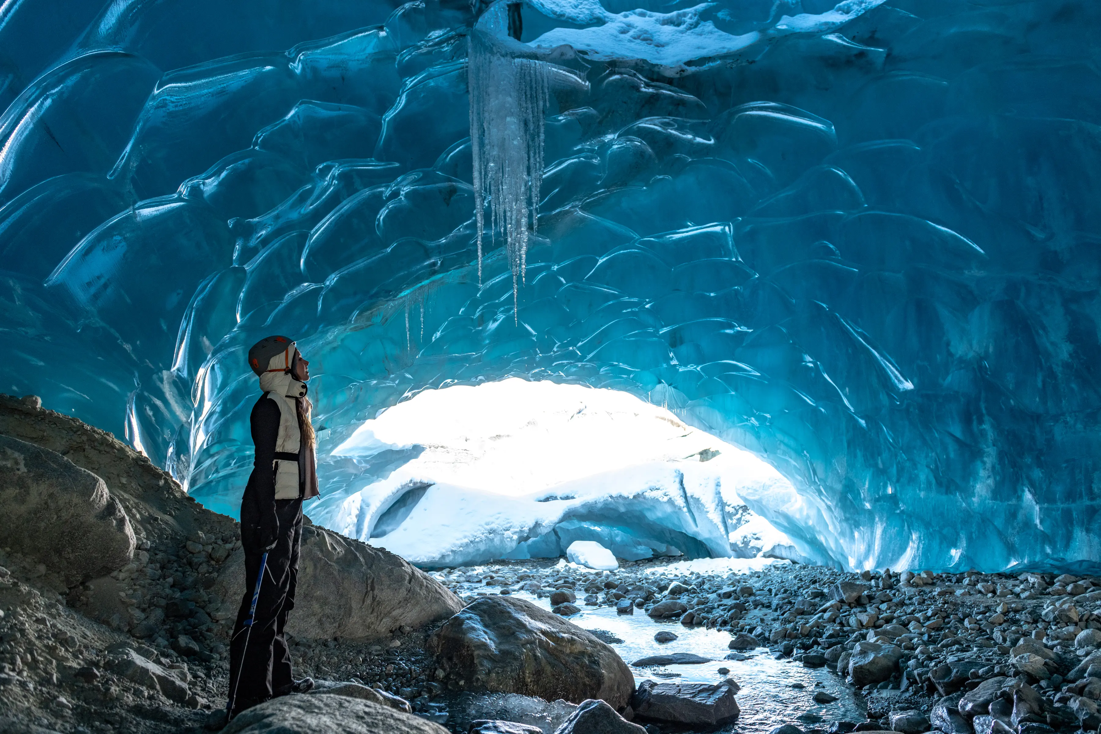 Person in winter gear standing inside an ice cave with icicles hanging from the ceiling and rocky ground.