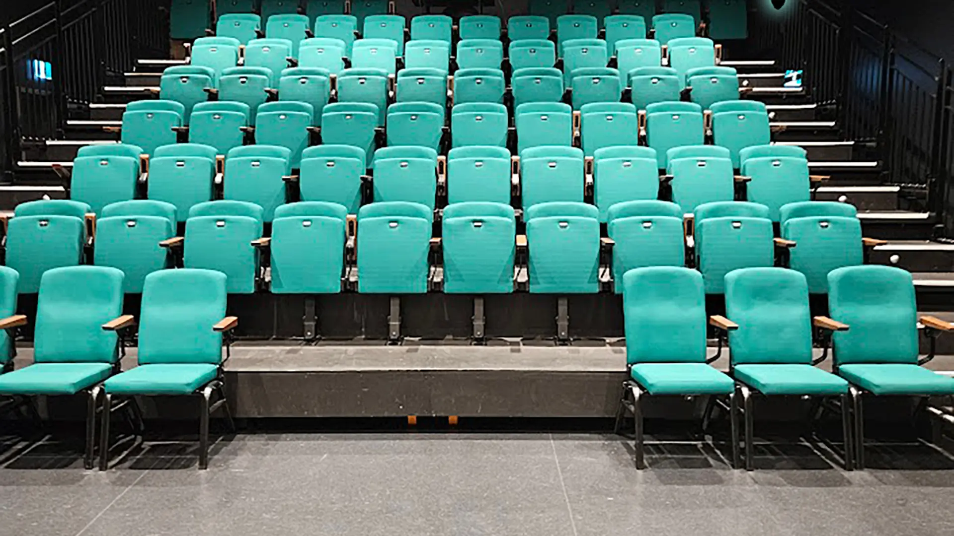 Empty auditorium with rows of teal cushioned seats and black metal frames.
