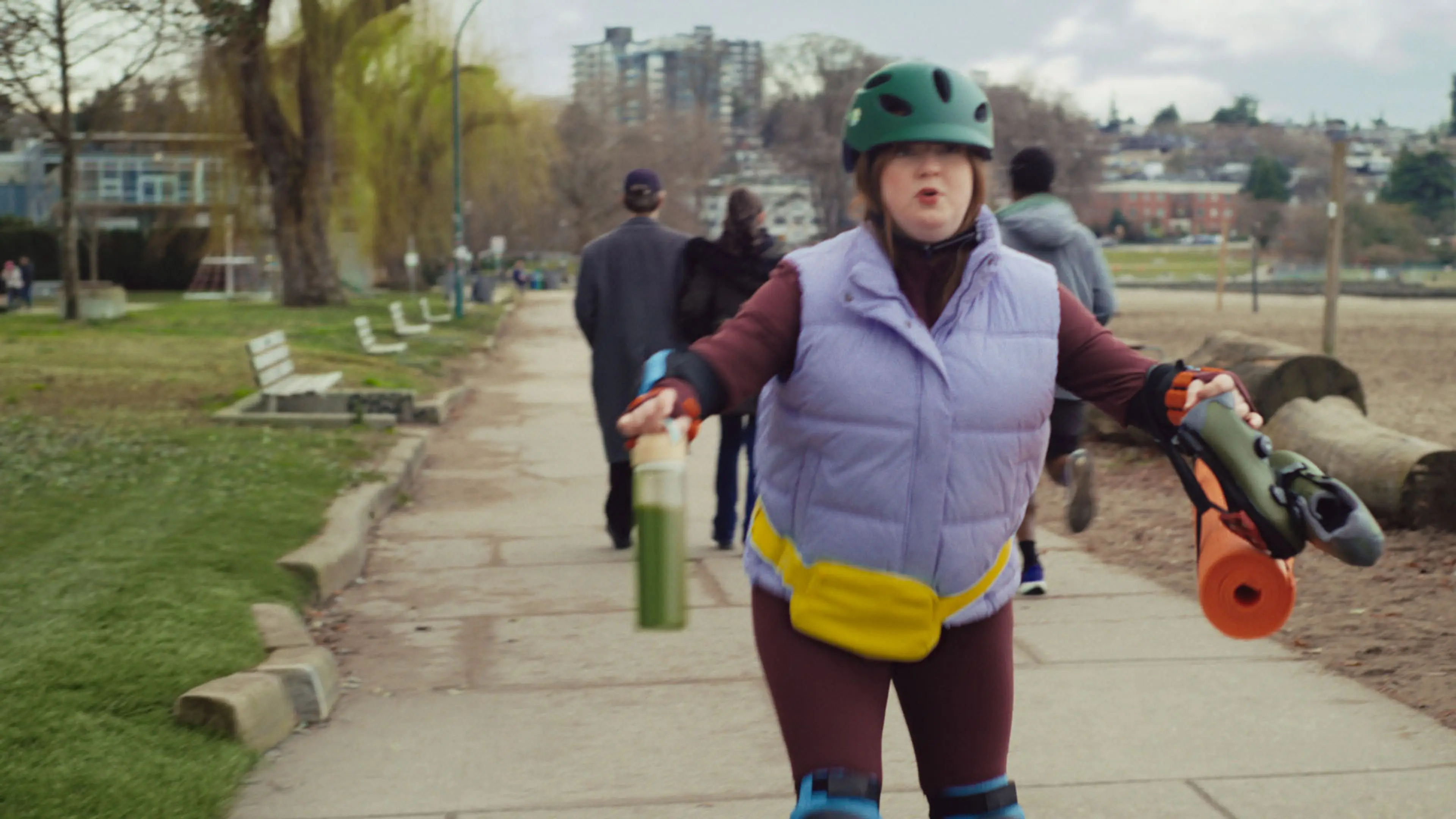 Person wearing a green helmet and purple vest holding a green drink and rollerblades on a park path.
