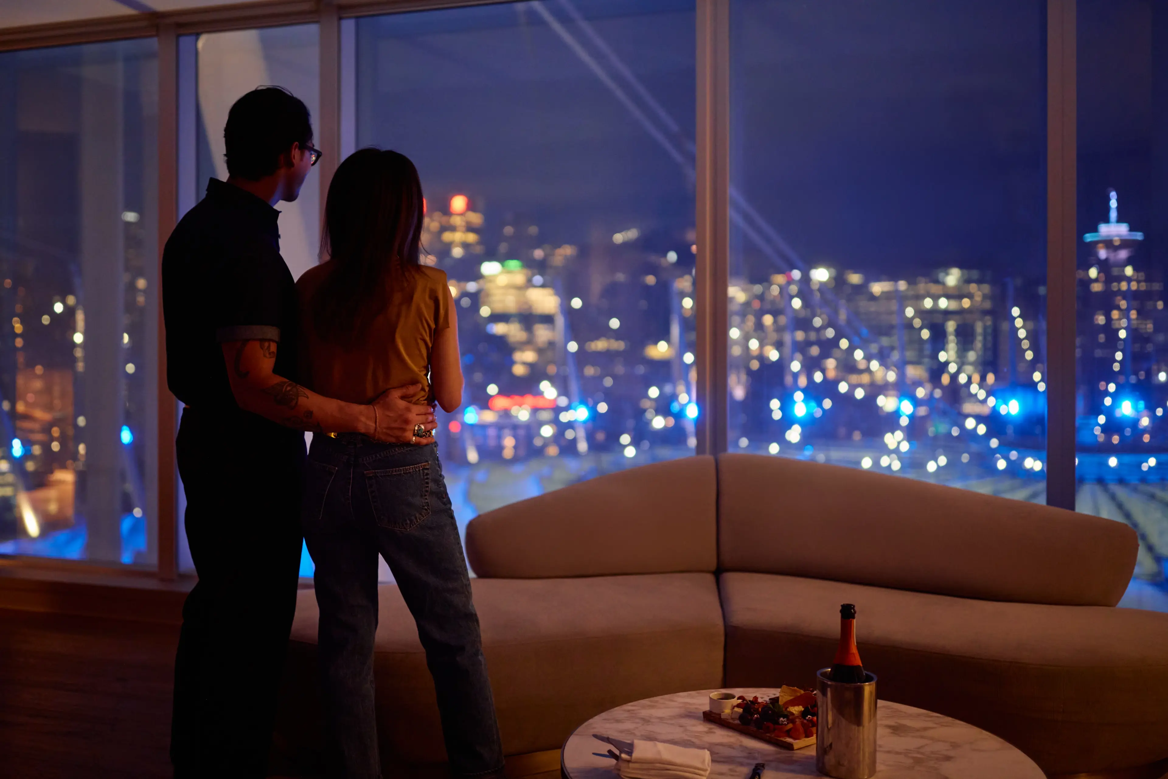A couple standing at the windows of the JW Marriott Parq Hotel looking towards BC Place lit up at dusk.
