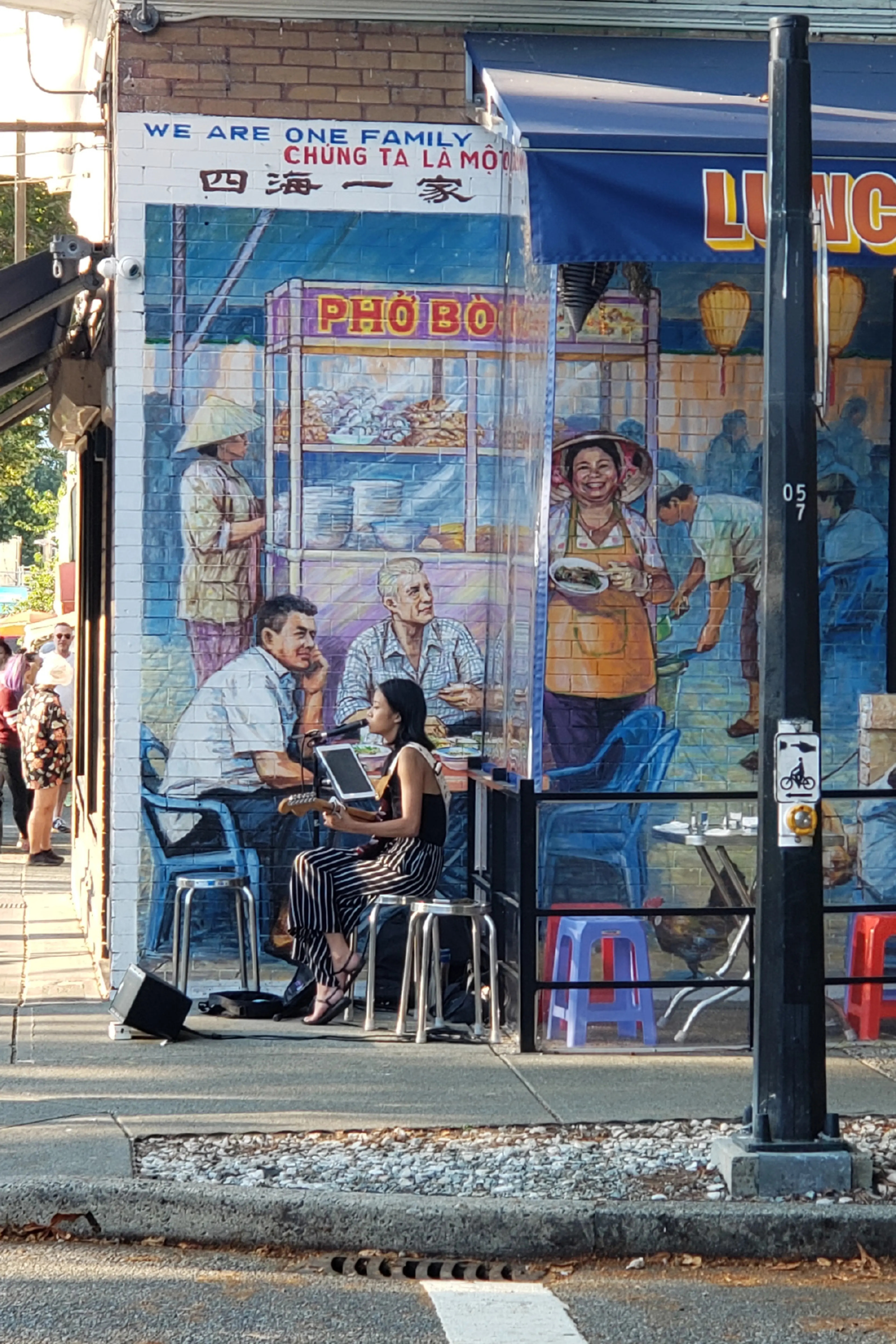 Woman playing guitar and singing outside a restaurant with a mural of people eating and serving pho on the wall.