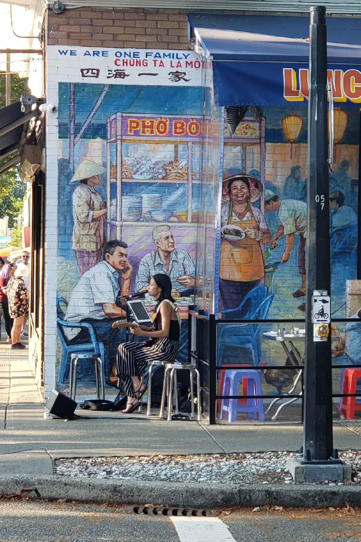 Woman playing guitar and singing outside a restaurant with a mural of people eating and serving pho on the wall.
