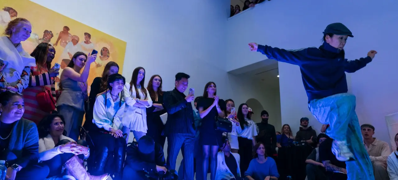 A person in a cap and blue outfit dancing energetically in front of a seated and standing audience in a gallery-like space with blue lighting.