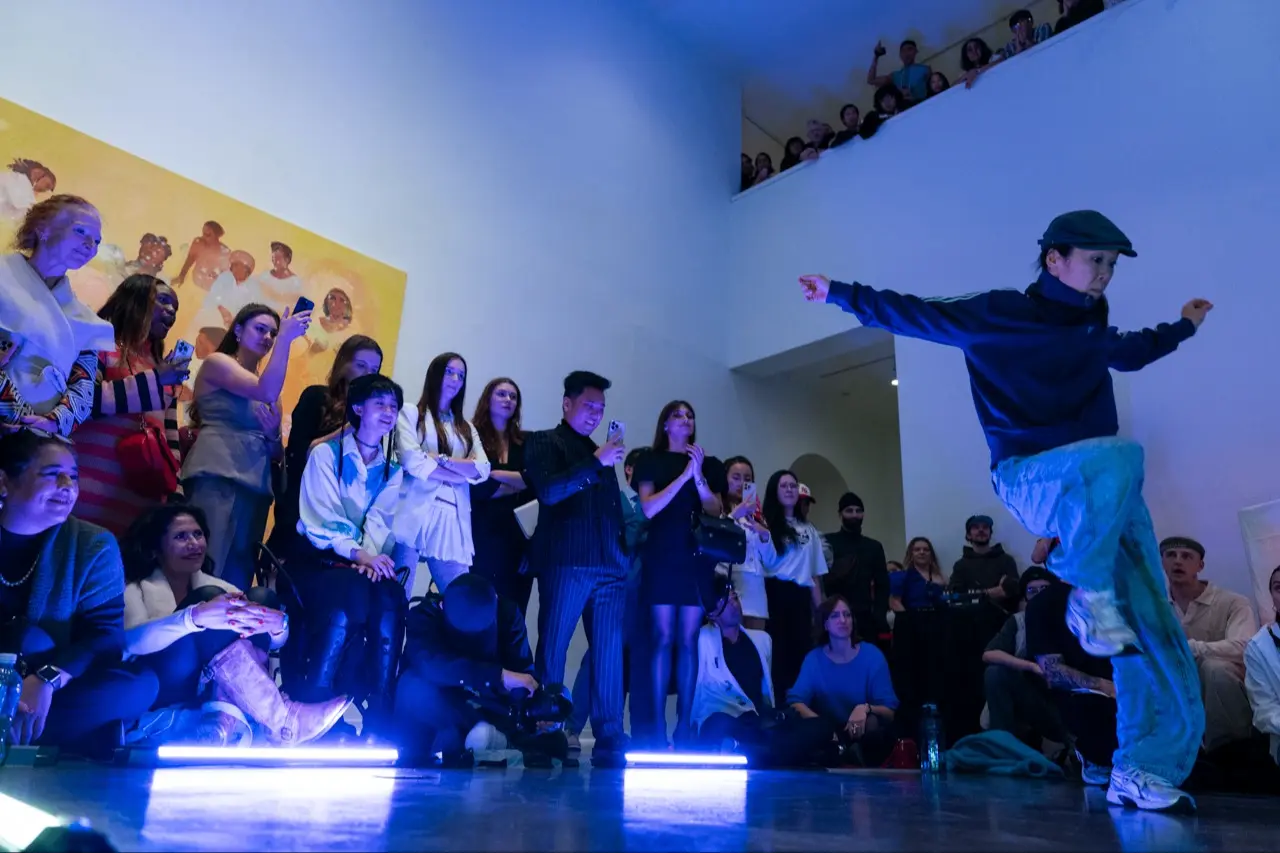 A person in a cap and blue outfit dancing energetically in front of a seated and standing audience in a gallery-like space with blue lighting.