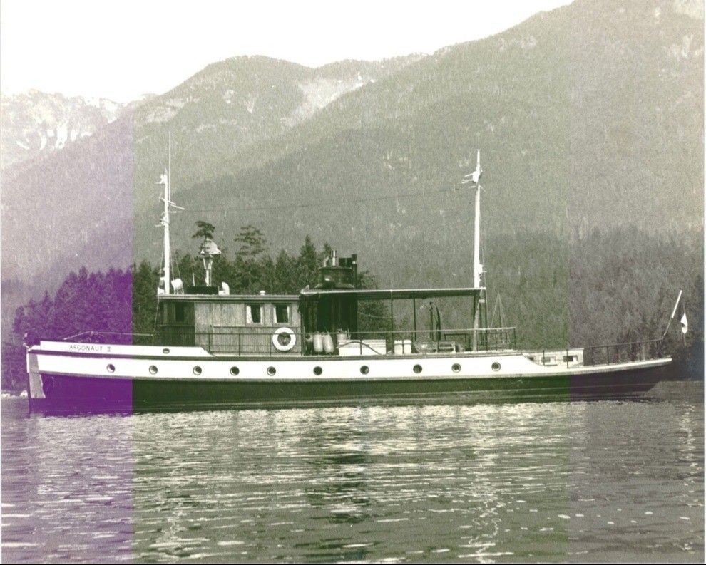 Side view of a vintage boat named Argonaut II on calm water with forested mountains in the background.