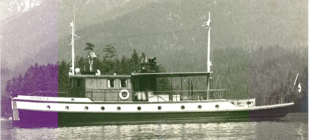 Side view of a vintage boat named Argonaut II on calm water with forested mountains in the background.