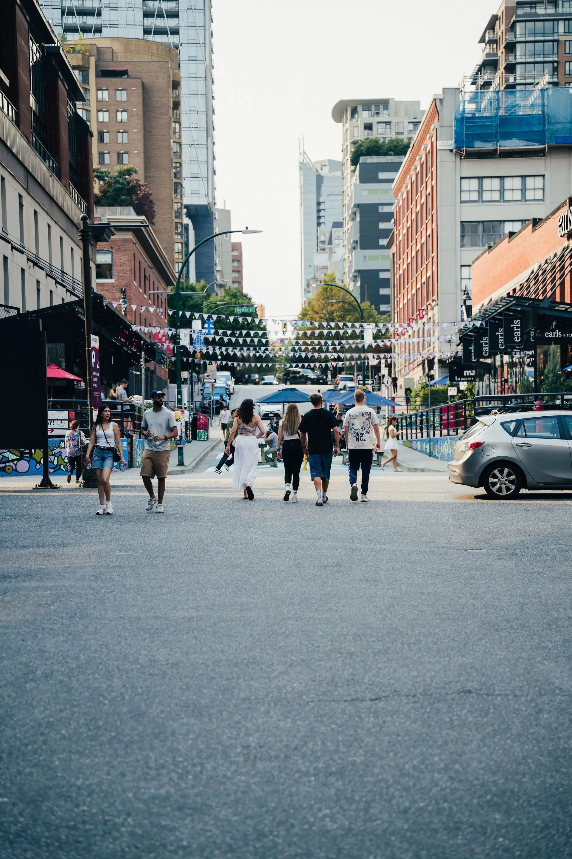 People walking across a city street with buildings, cars, and decorative flags overhead.