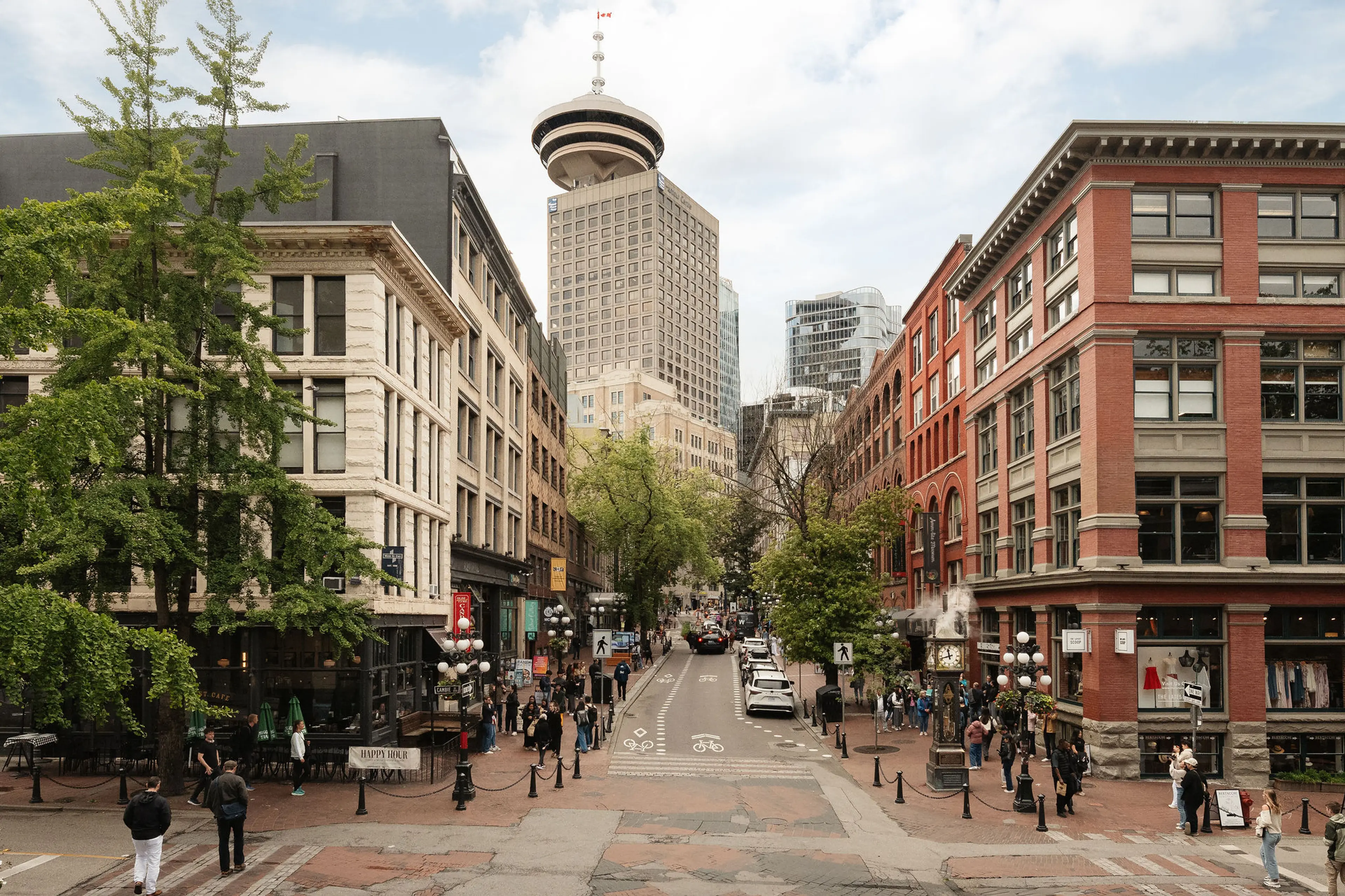 Urban street scene with pedestrians, historic buildings, trees, and a tall tower with a Canadian flag in the background.