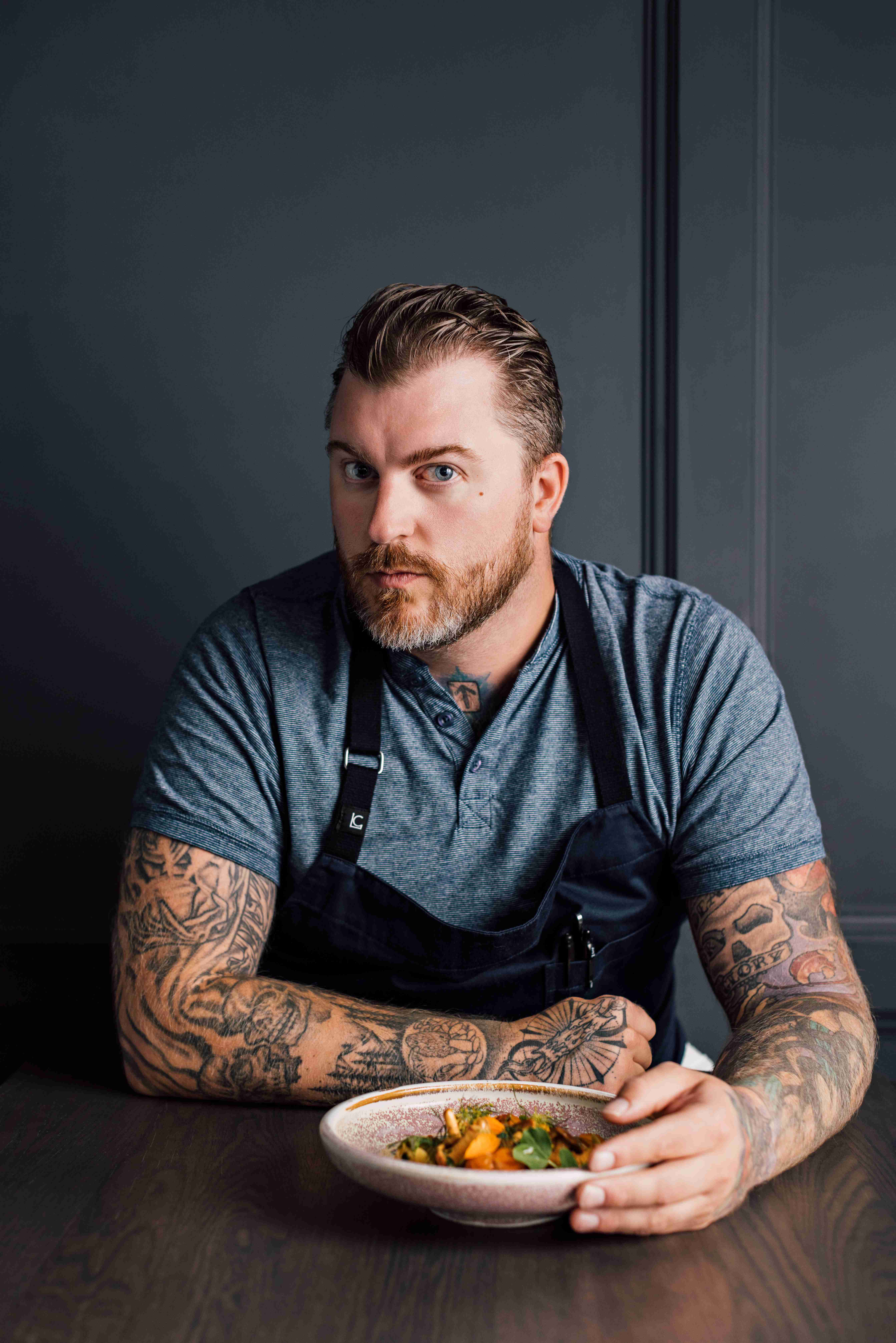 Man with tattoos wearing a gray shirt and black apron holding a bowl of food at a wooden table.
