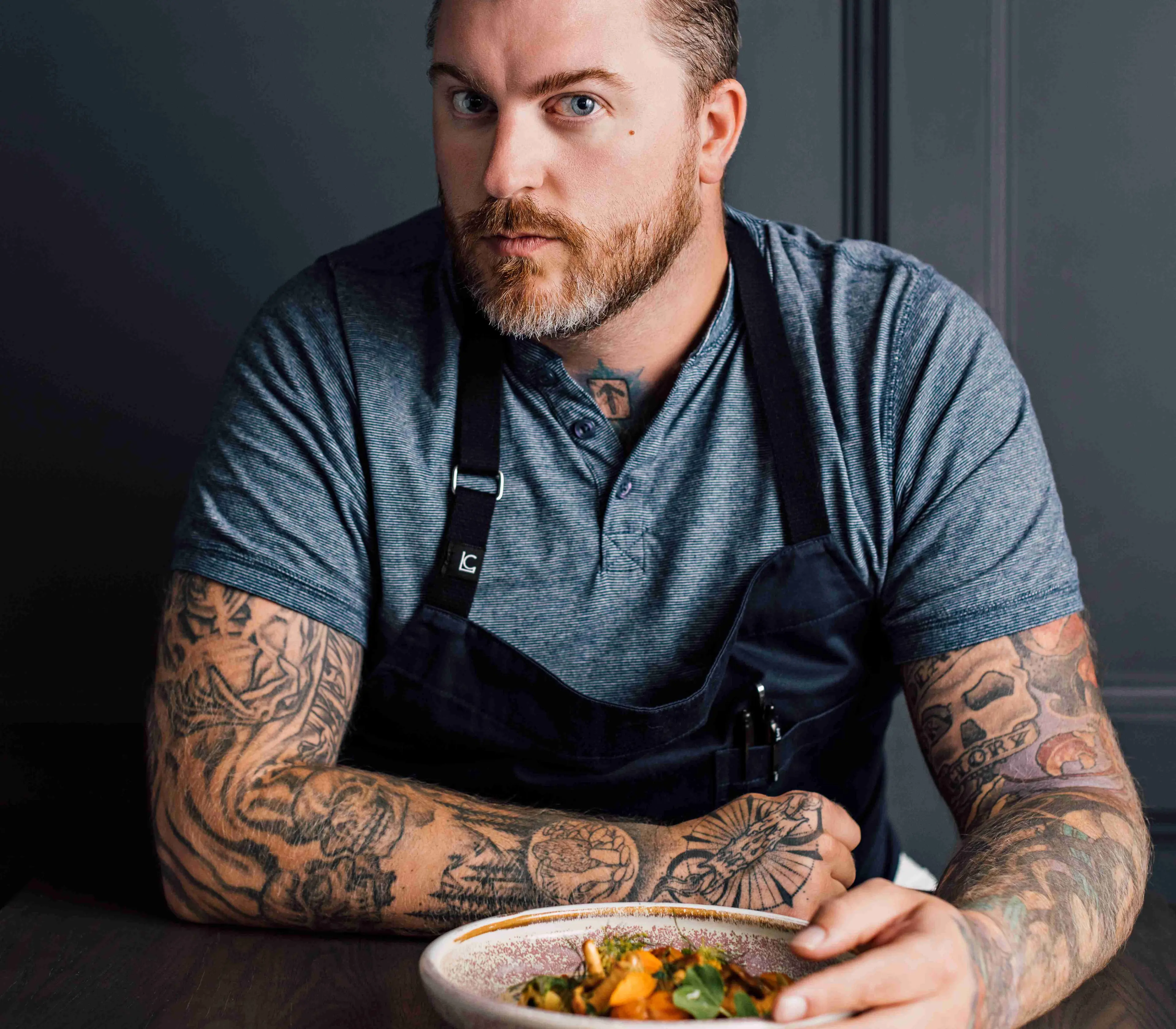Man with tattoos wearing a gray shirt and black apron holding a bowl of food at a wooden table.