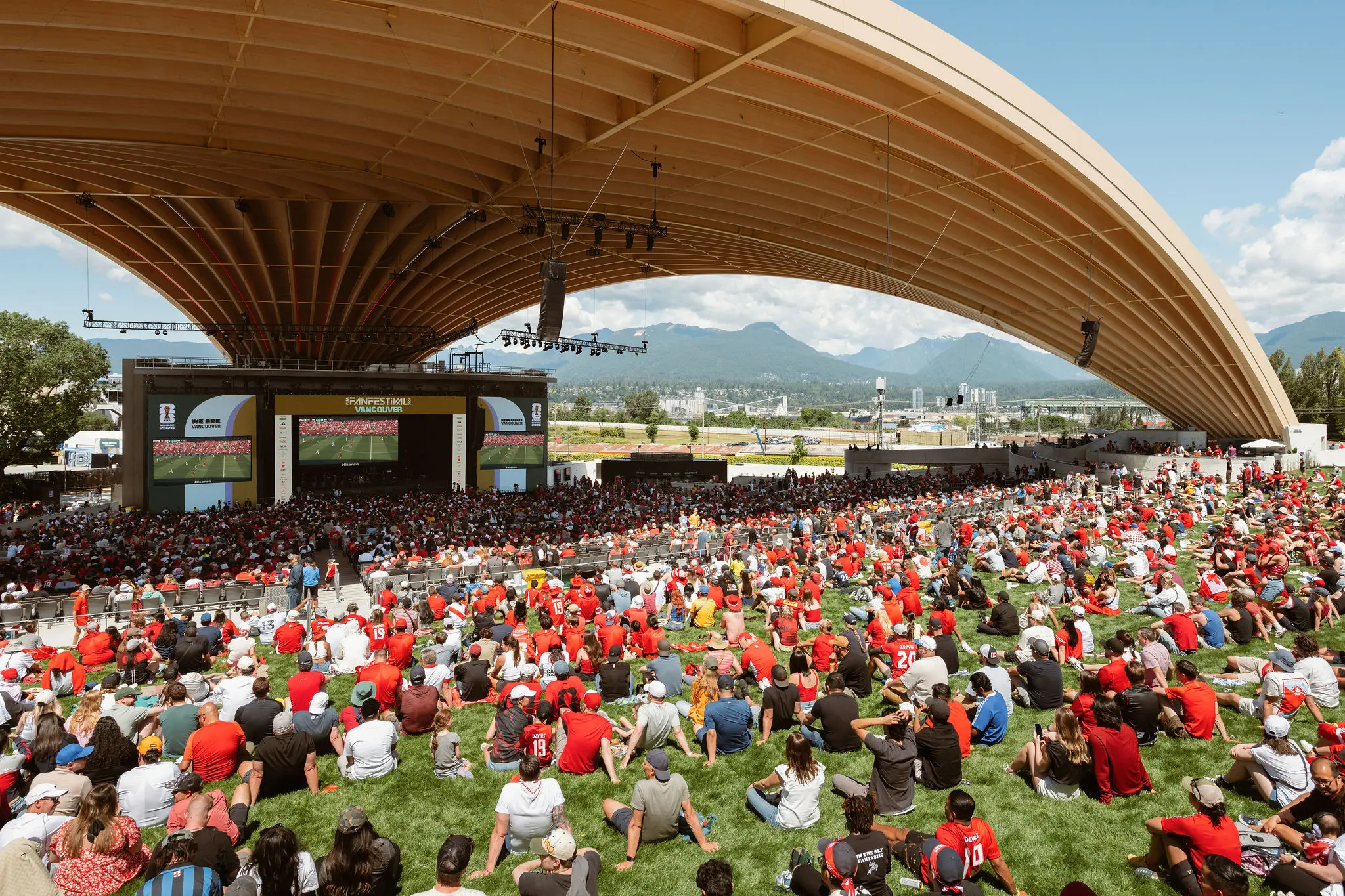 Large crowd of people sitting on grass and seats under a large curved roof watching a sports game on big screens outdoors with mountains in the background.