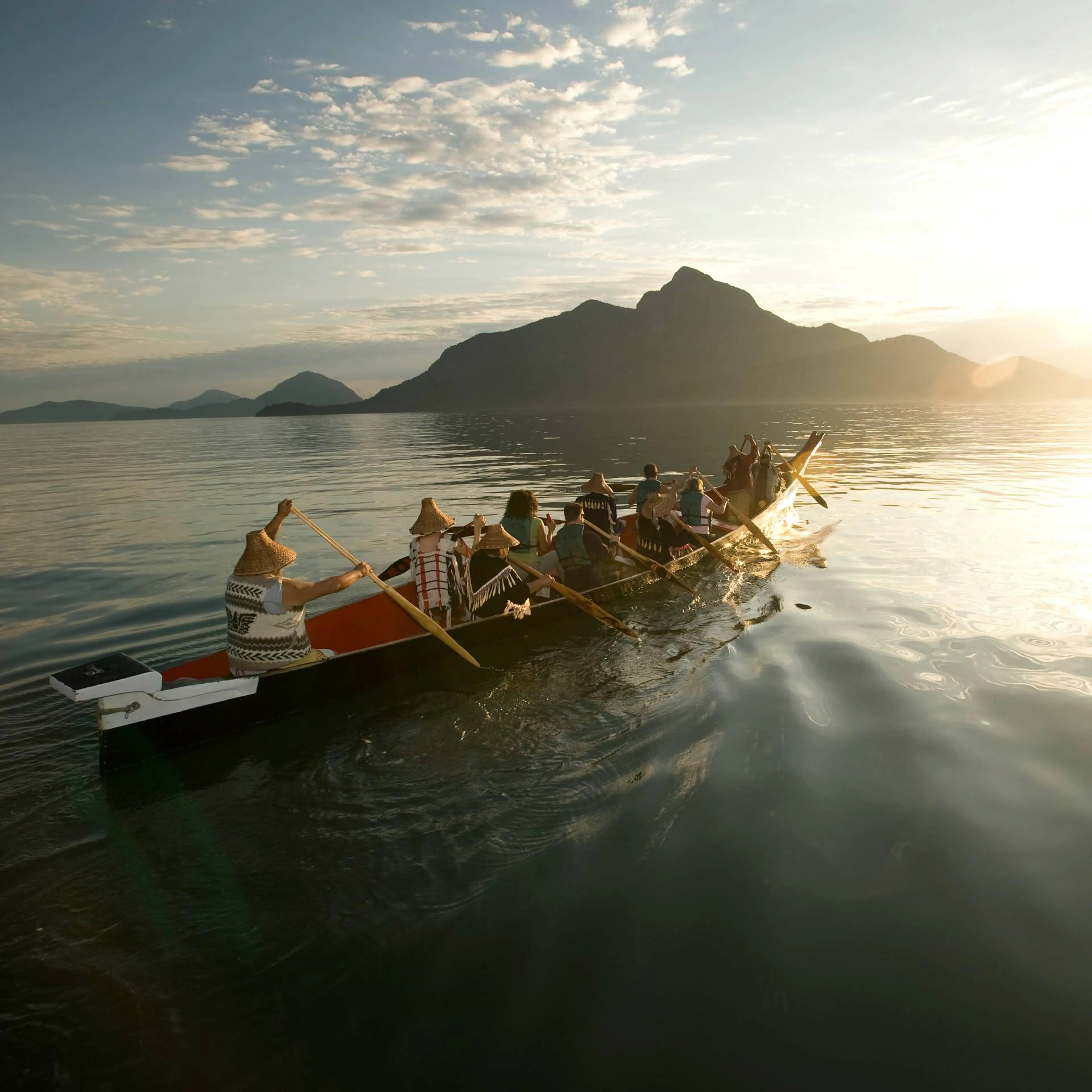 Group of people paddling a long canoe on calm water with mountains and a cloudy sky at sunset.