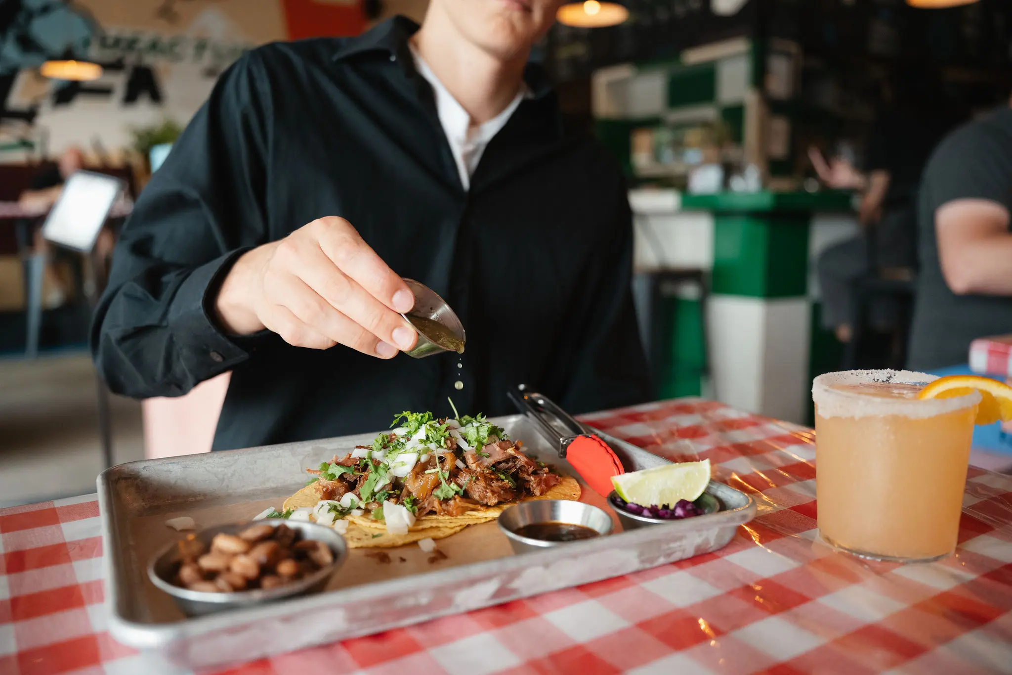 Person in black shirt squeezing lime juice over tacos on a metal tray with sides and a drink on a red and white checkered tablecloth.